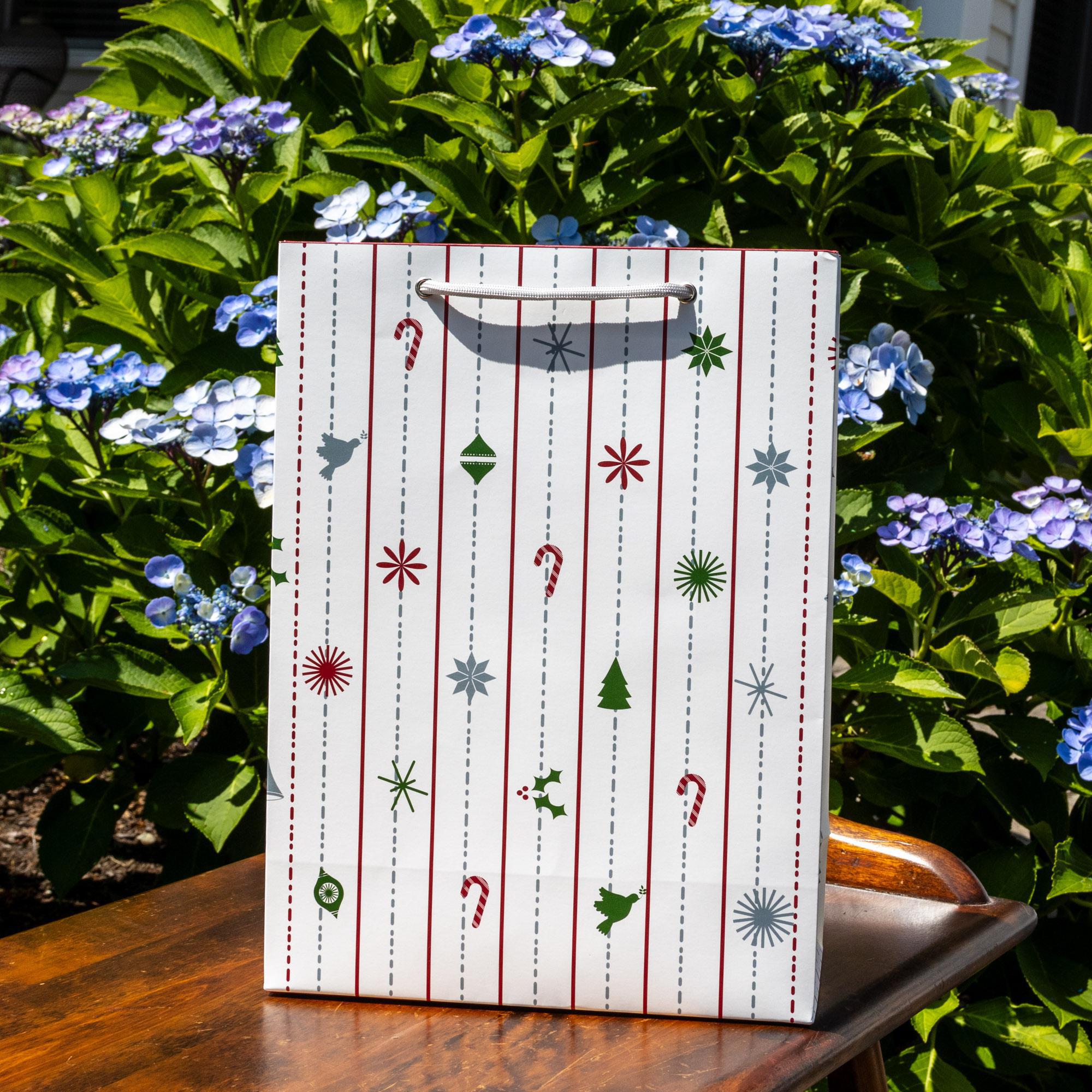 White Christmas gift bag with vertical red stripes and festive icons including candy canes, trees, and ornaments, displayed outdoors on a wooden surface surrounded by vibrant blue hydrangea flowers and lush green foliage