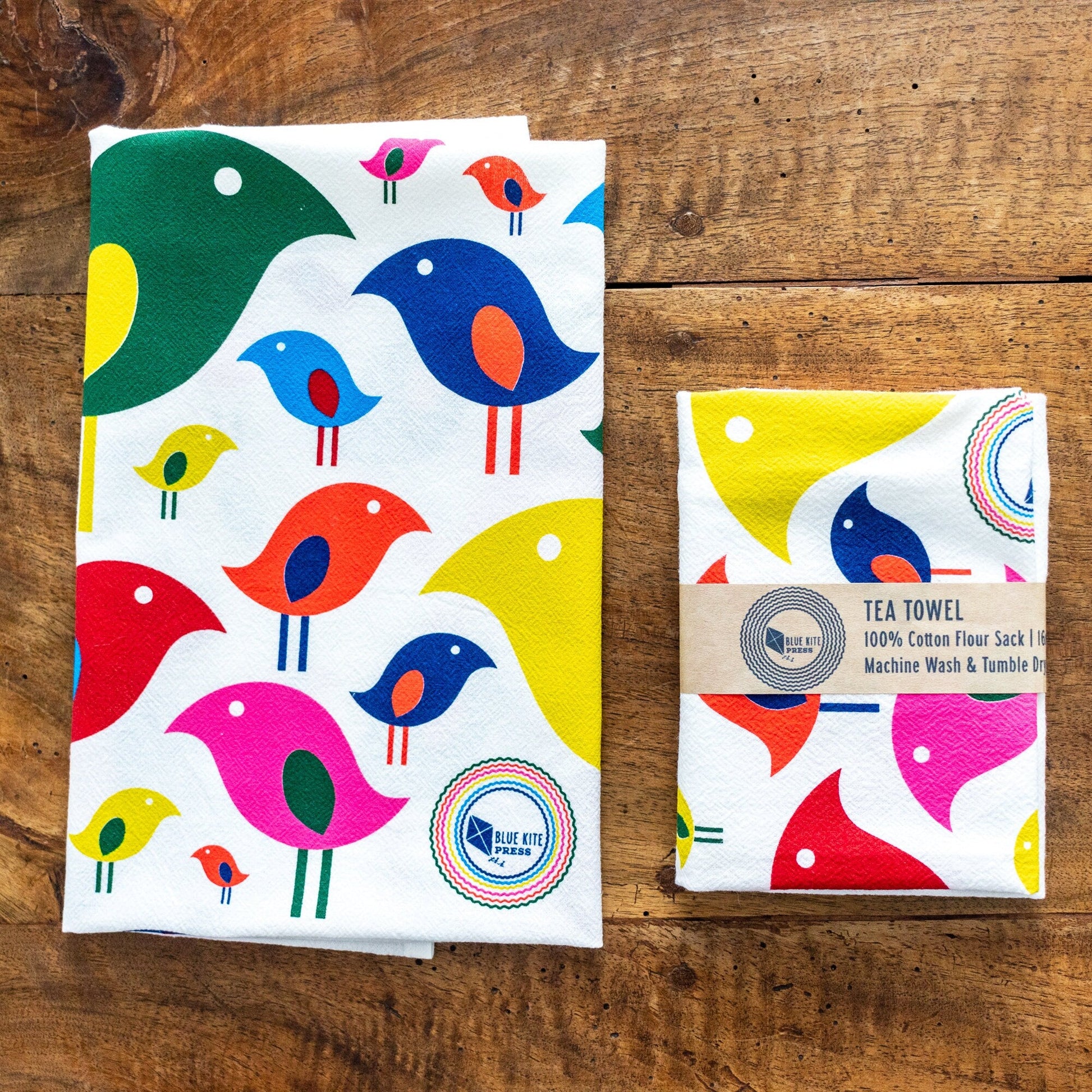 Colorful mid-century inspired bird pattern tea towels made from 100% cotton flour sack, displayed unfolded and folded on wooden surface. Vibrant, stylized birds in various colors and sizes create a cheerful, retro design perfect for modern kitchens.