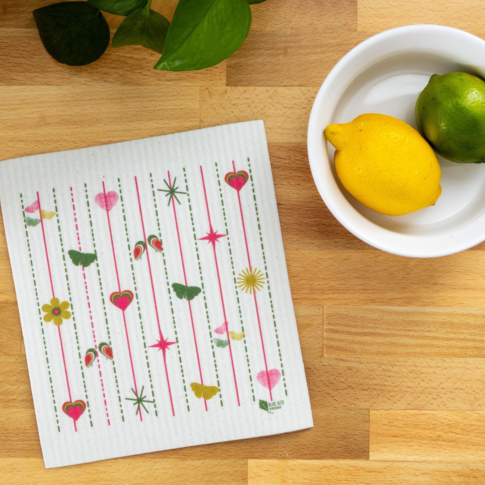 Pink and green Swedish dishcloth with hearts and flowers pattern on wooden table, next to white bowl with lemon and lime, and green leaves in corner