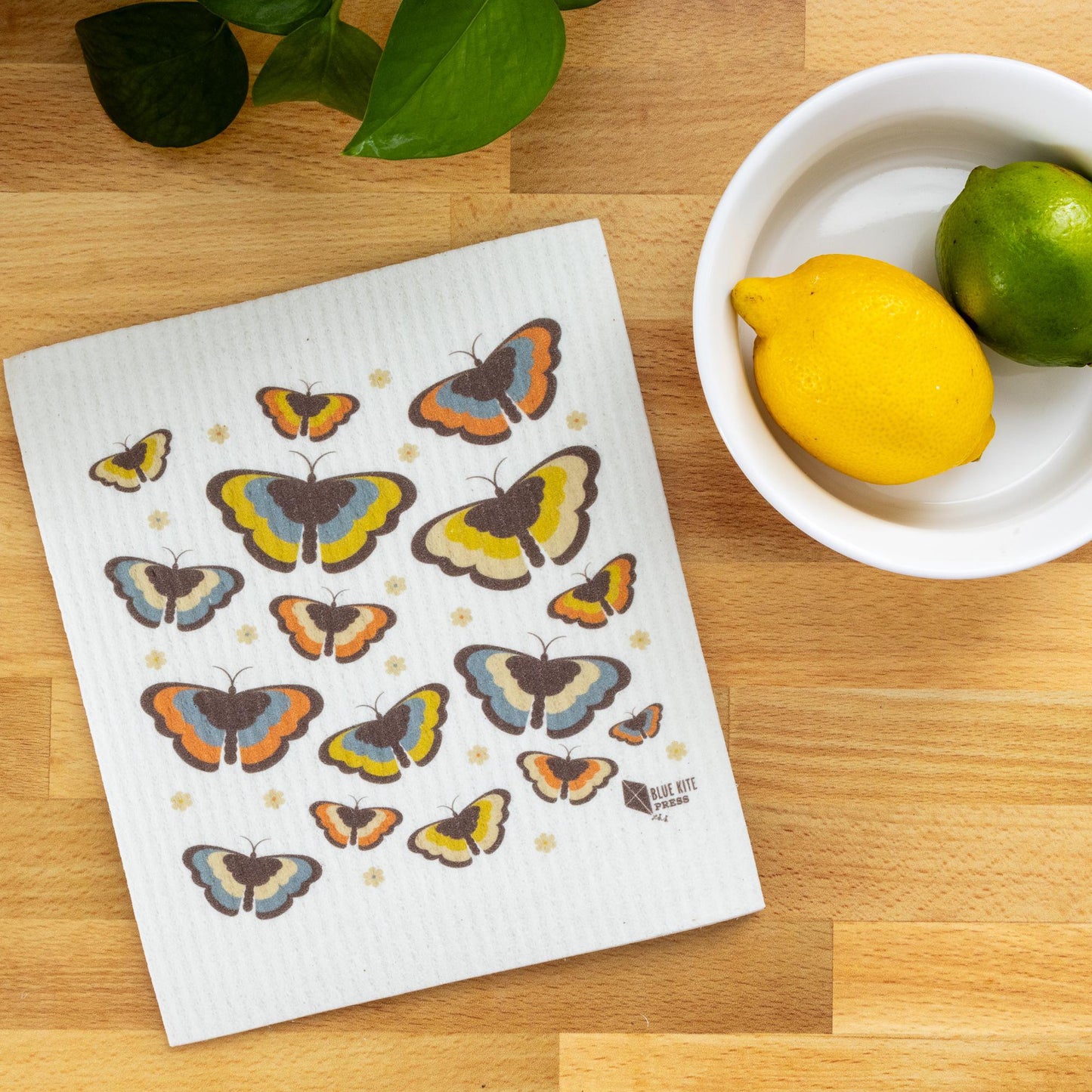 Butterfly Swedish dishcloth with 70s-inspired design on wooden surface, next to bowl of citrus fruits and green leaves, showcasing eco-friendly kitchen accessory and sustainable living