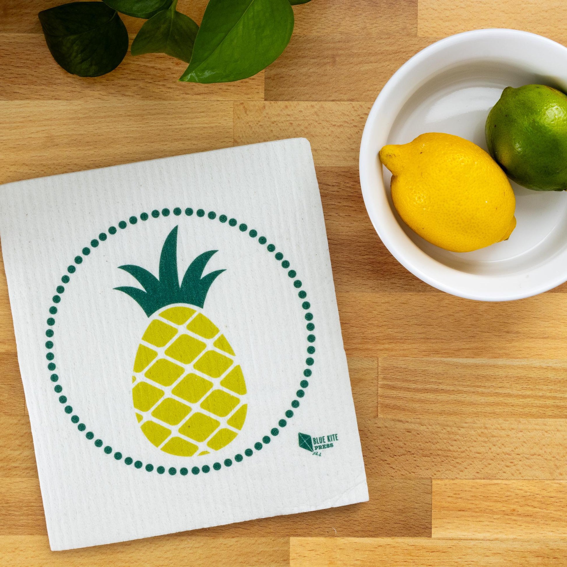 Pineapple Swedish dishcloth with modern yellow design on wooden surface, next to white bowl with lemon and lime, and green leaves for eco-friendly kitchen decor