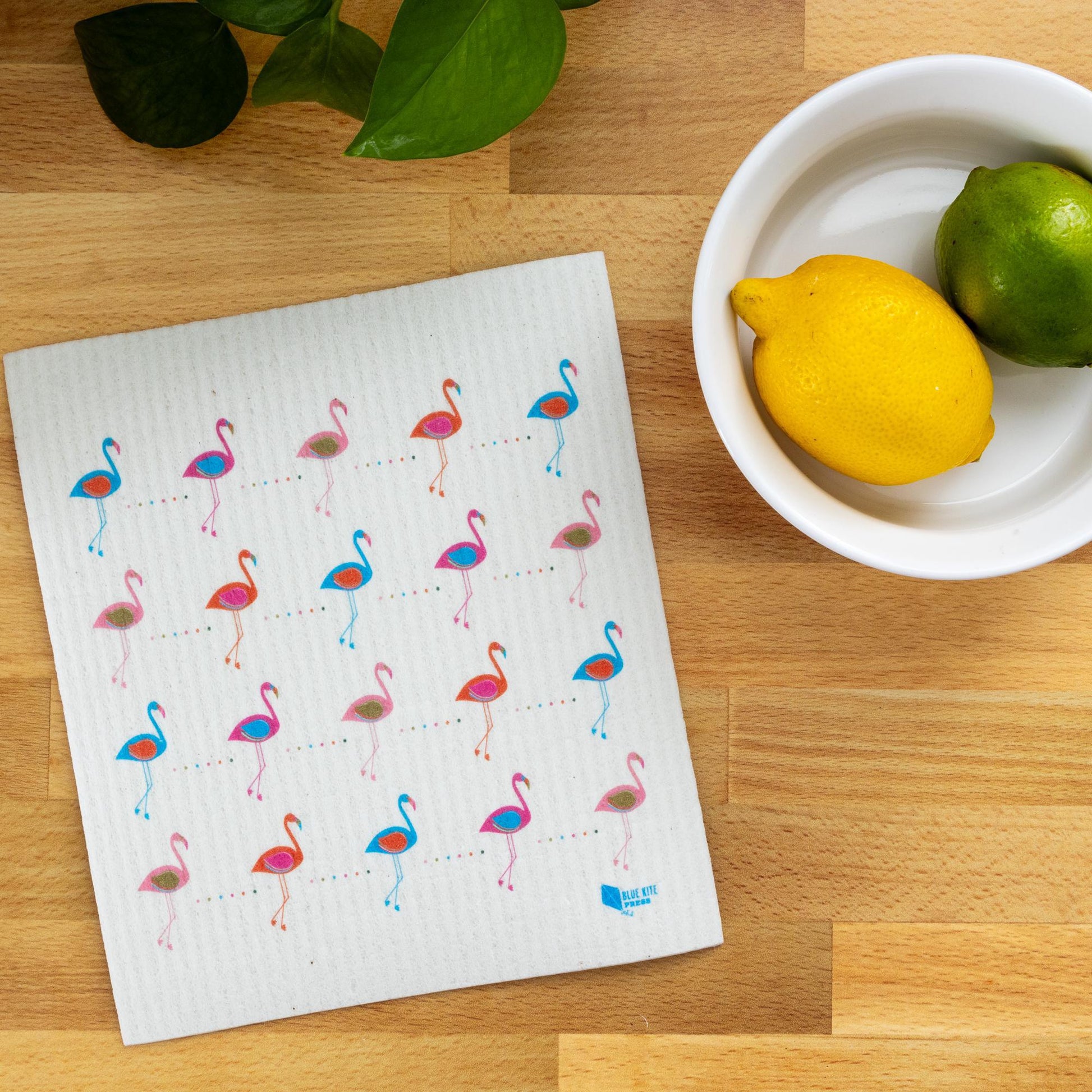 Swedish dishcloth with colorful flamingo pattern displayed on wooden countertop next to bowl of citrus fruits and green leaves, showcasing eco-friendly kitchen product design and functionality