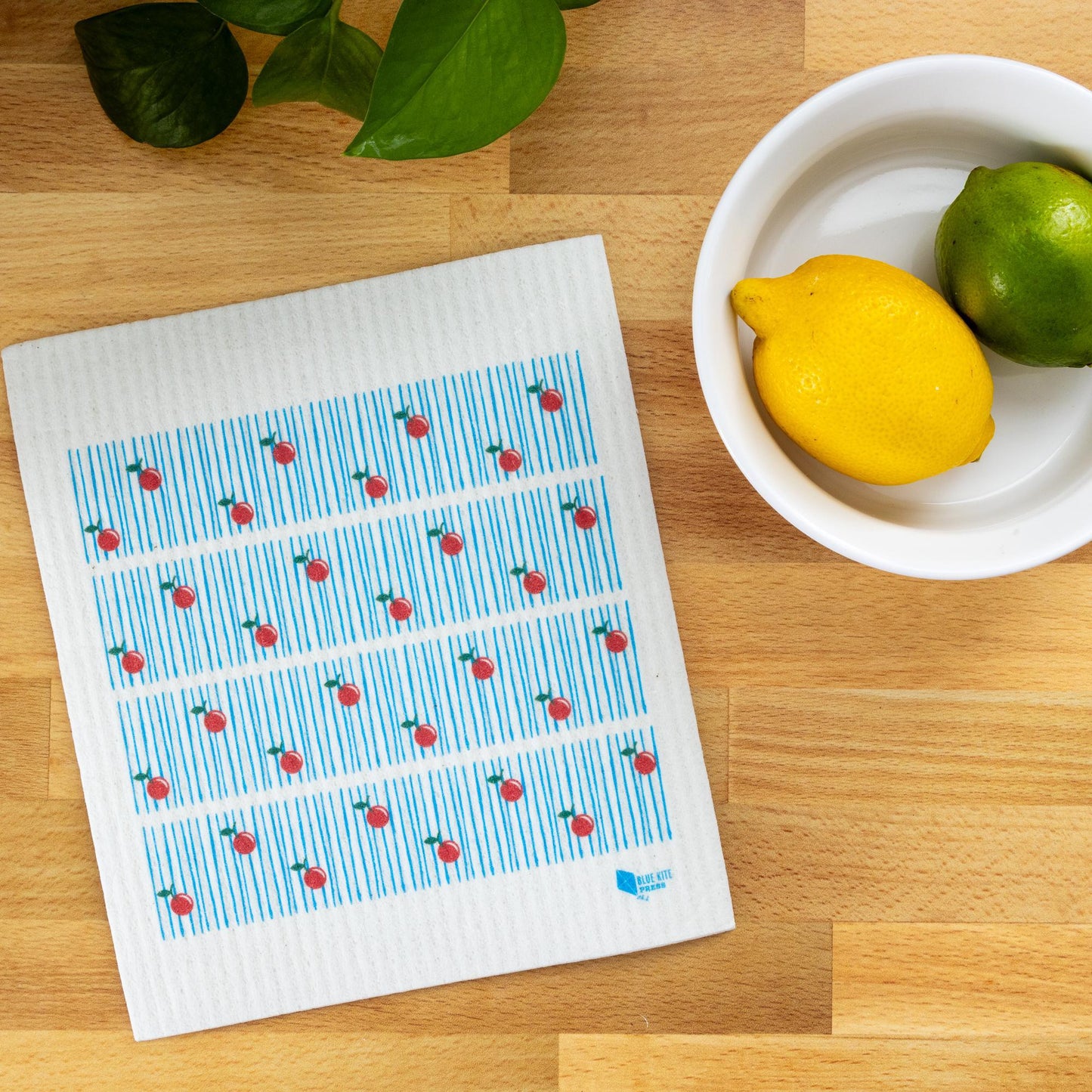 Swedish dishcloth with red cherry pattern on blue stripes, displayed on wooden surface next to white bowl containing lemon and lime, with green leaves in corner