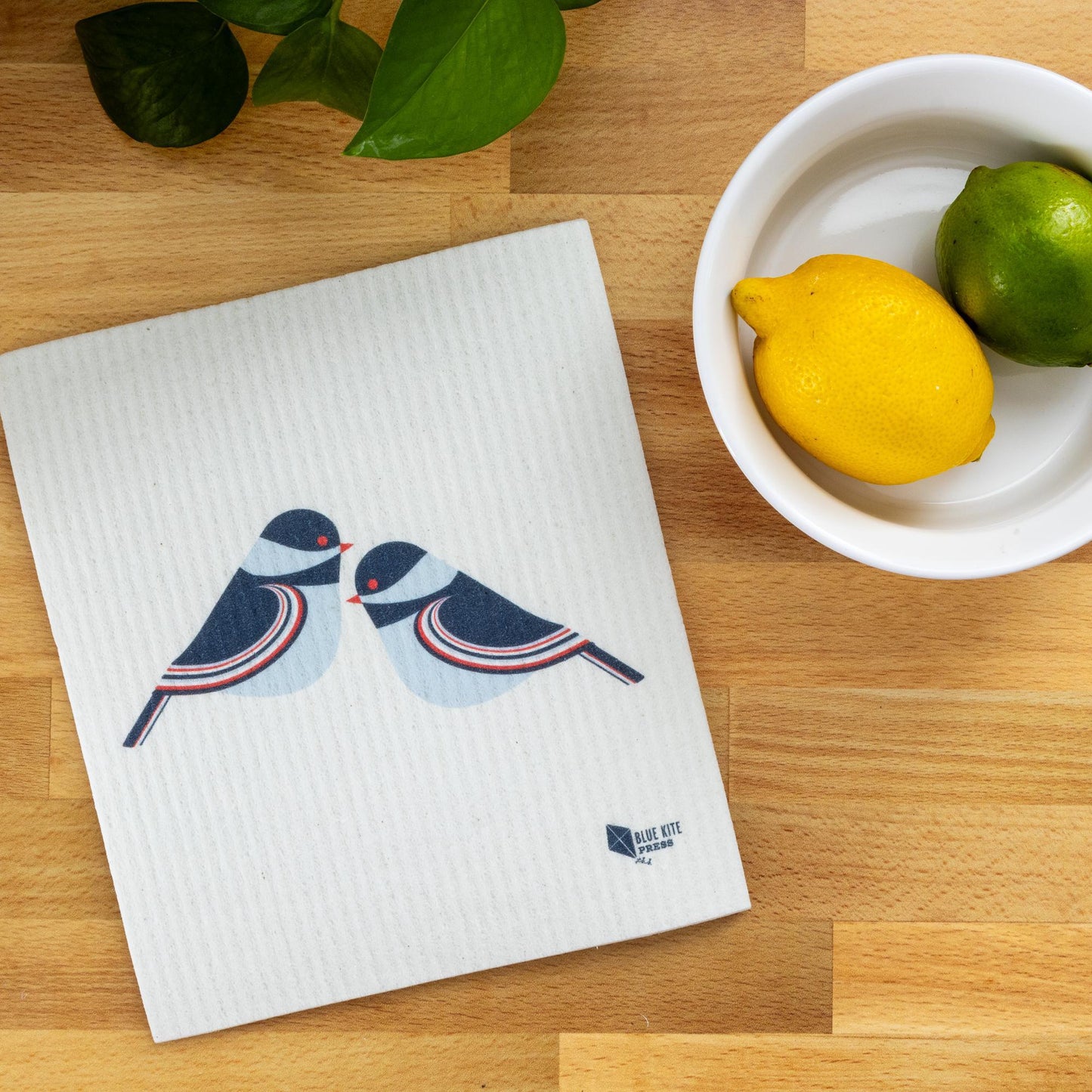 Eco-friendly Swedish dishcloth with modern chickadee design, displayed on wooden surface next to bowl of citrus fruits and green leaves, showcasing sustainable kitchen cleaning alternative