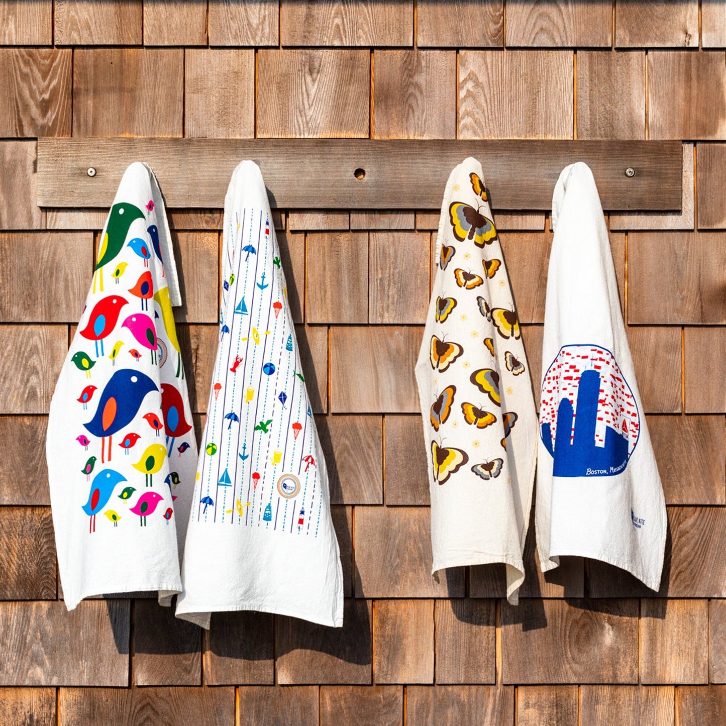 Four white flour sack towels hanging on wooden wall, featuring colorful modern bird flock, butterfly, and abstract designs, showcasing the Bright Bird Flour Sack Towel product in a rustic setting