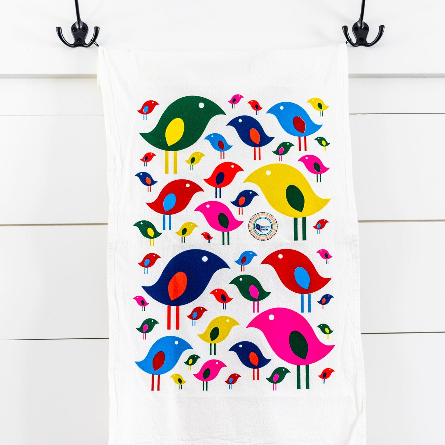 Colorful mid-century inspired bird pattern tea towel hanging on hooks, featuring vibrant stylized birds in various sizes and colors on white cotton fabric, showcasing a playful and retro kitchen accessory design