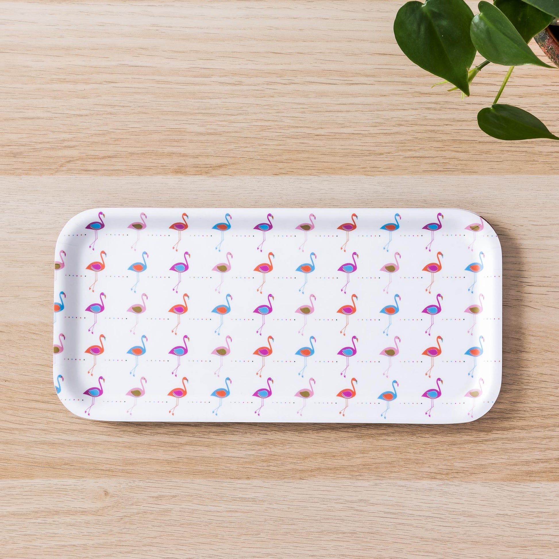 Rectangular birch tray with repeating pattern of pink and blue flamingos on white background, displayed on light wooden surface with green plant leaves visible