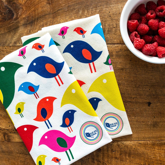 Colorful bird-patterned tea towels on wooden surface next to bowl of fresh raspberries, showcasing vibrant mid-century inspired design and eco-friendly cotton material for kitchen use