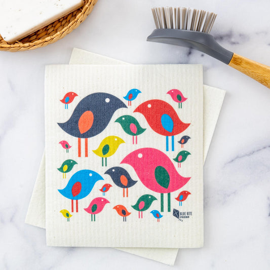 Bright Bird Swedish Dishcloth featuring colorful minimalist bird flock pattern in red, blue, navy, pink, green, and yellow on white cellulose background, displayed with cleaning brush on marble surface. Eco-friendly reusable kitchen cloth with modern Scandinavian design.