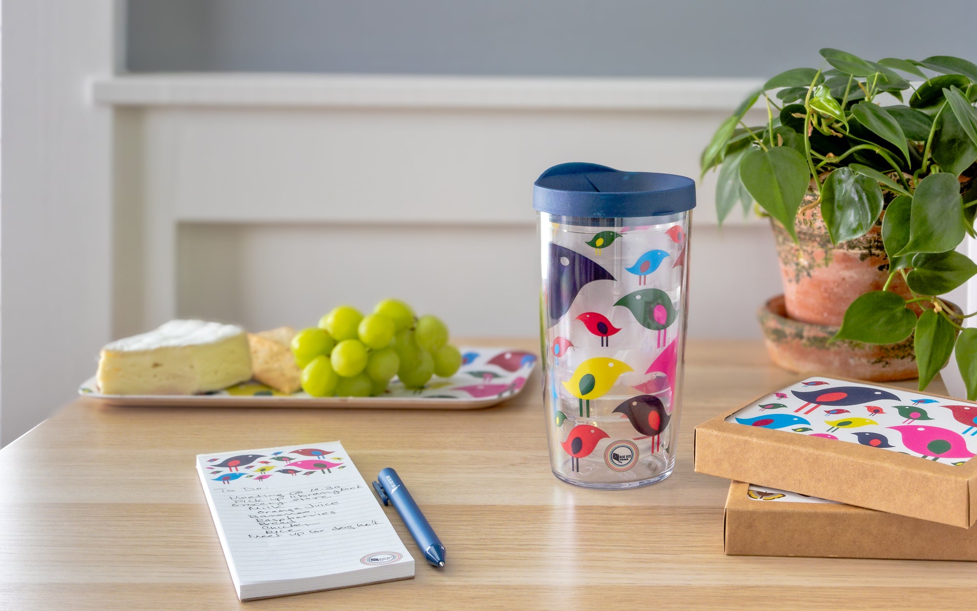 Colorful modern birds decorating a Tervis tumbler, notepad, notecard set and birch tray with cheese, grapes and a plant
