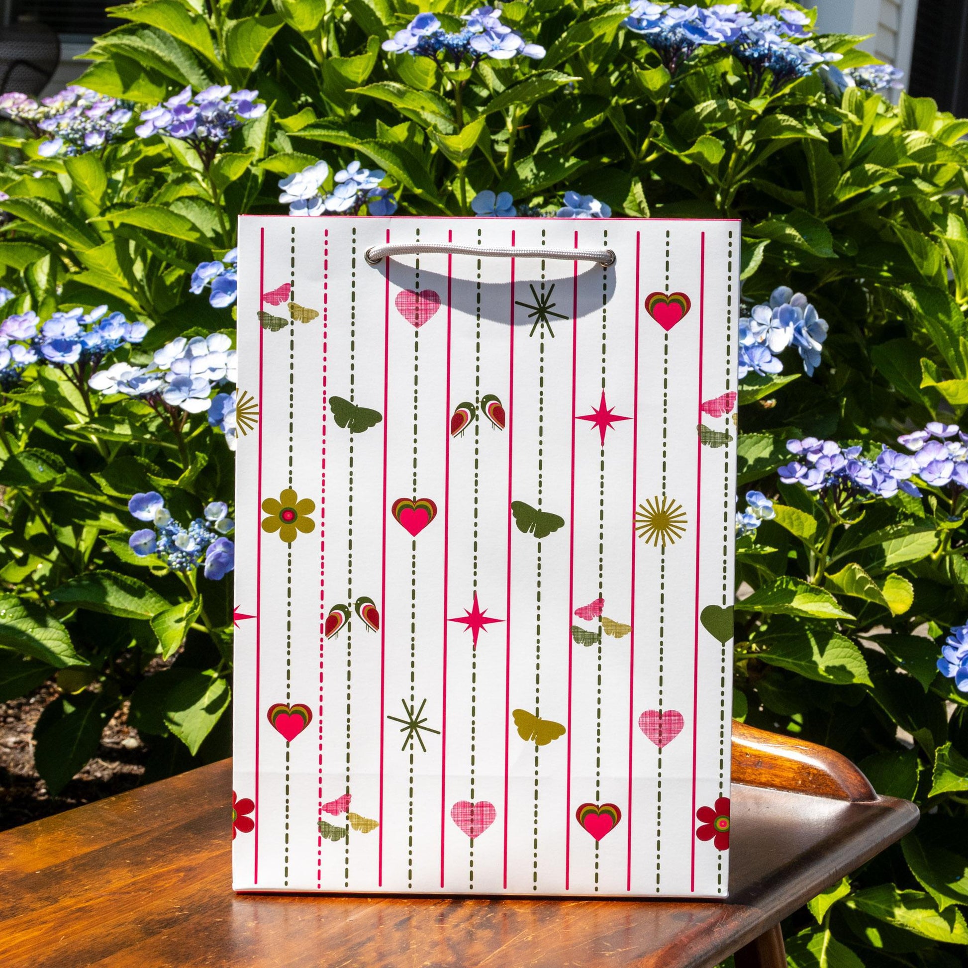 Modern pink and green gift bag with vertical dotted lines featuring hearts, flowers, butterflies, and stars pattern, displayed outdoors against vibrant blue hydrangeas and lush green foliage