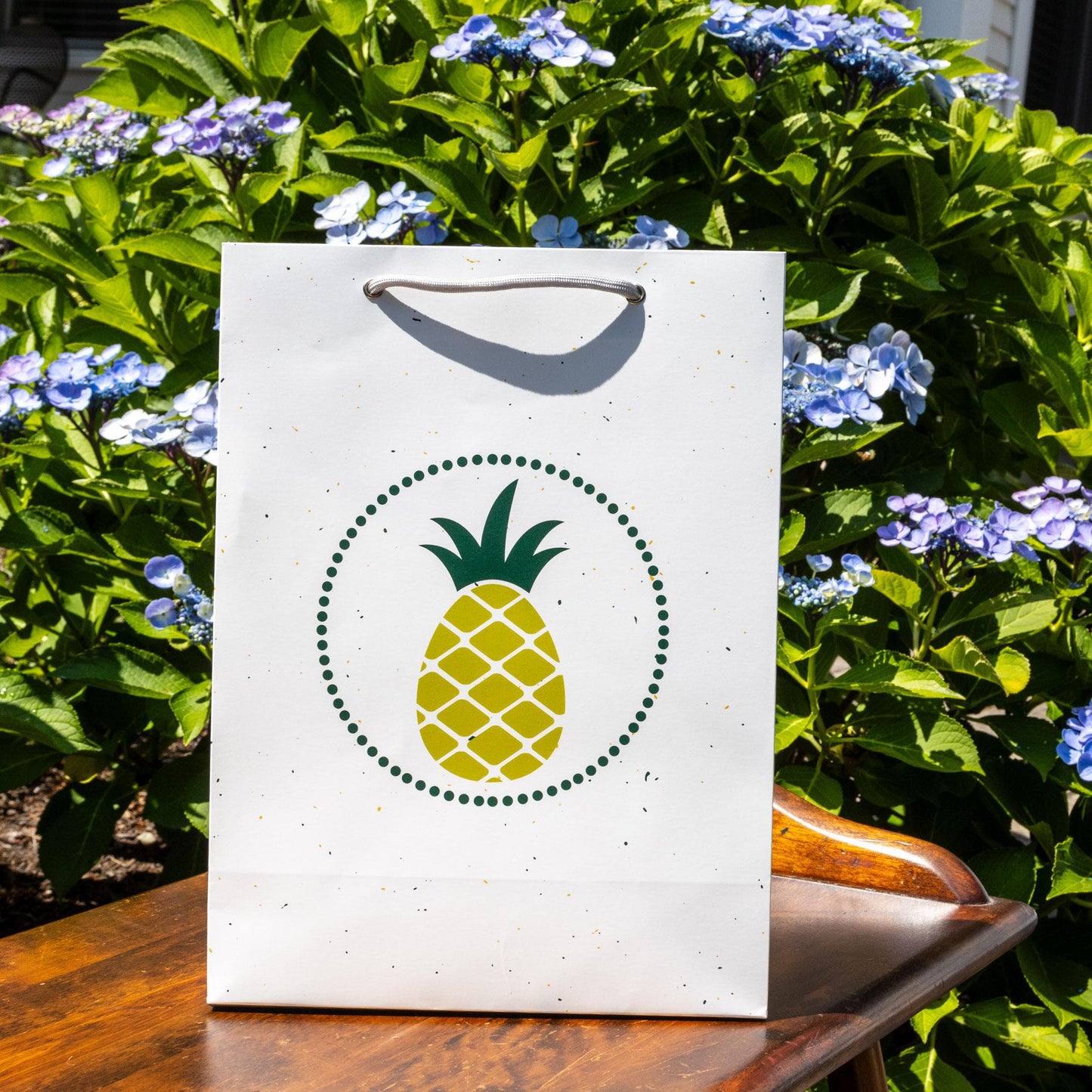 White gift bag with modern yellow pineapple design on front, surrounded by blue hydrangea flowers and green foliage, showcasing cheerful tropical theme for gift-giving