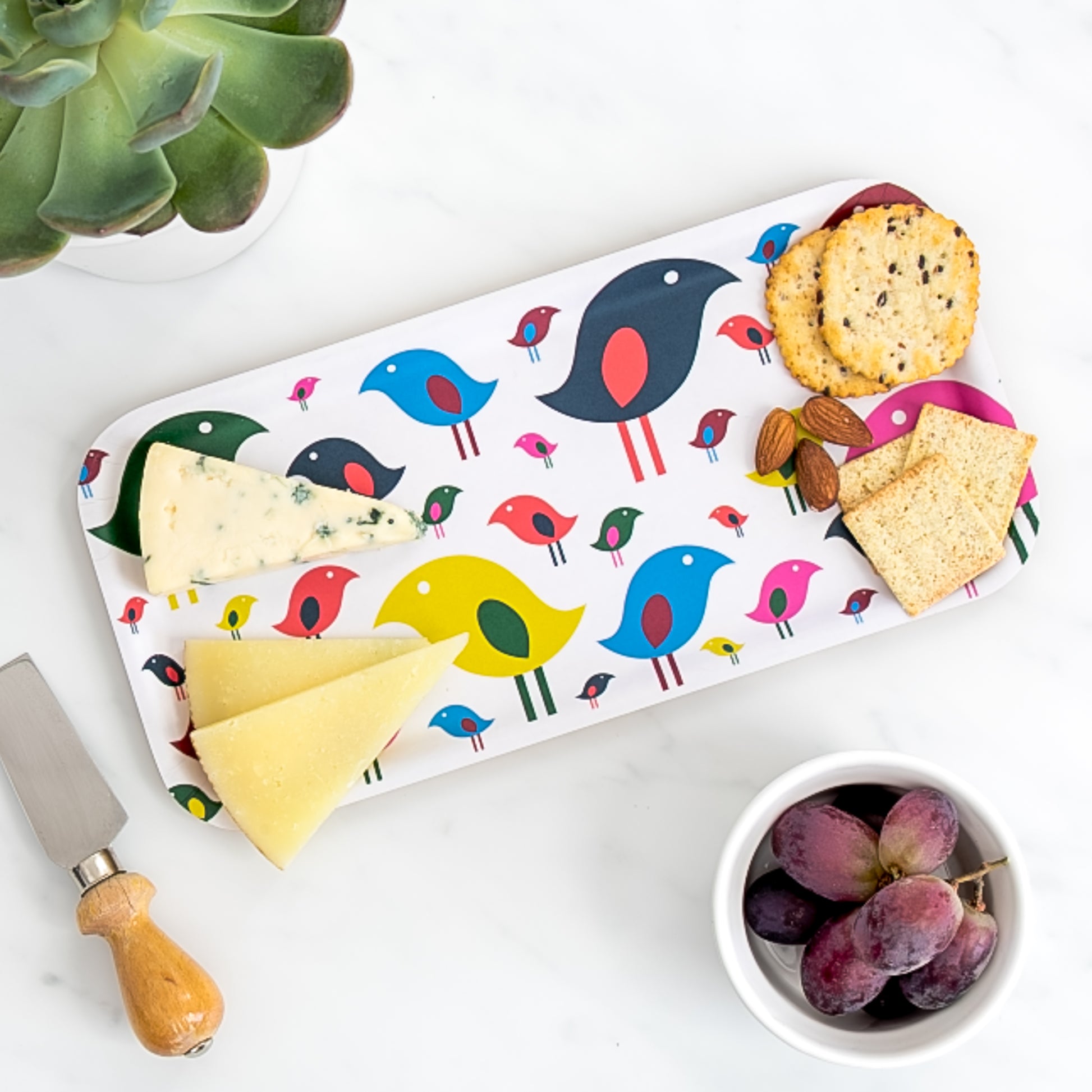 Cheese board of assorted cheese and crackers on birch tray with modern & joyful bright bird pattern