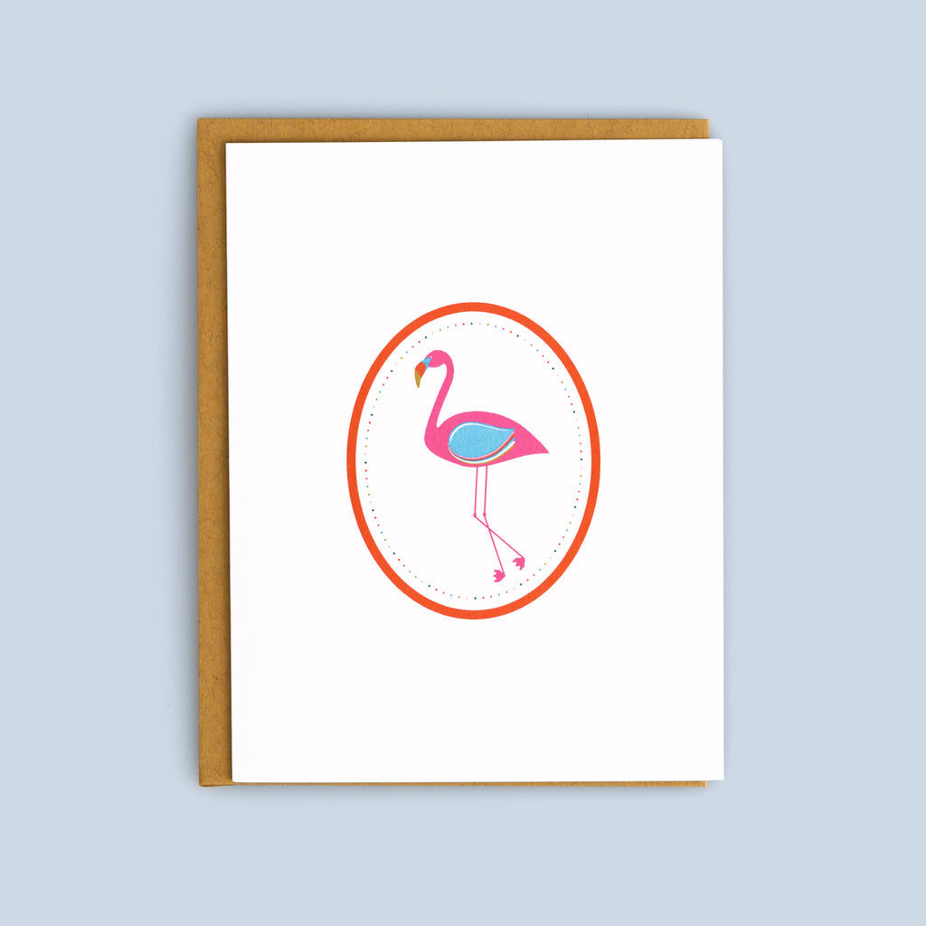 Flat card featuring a pink flamingo inside a red oval frame with a kraft envelope on a light blue background
