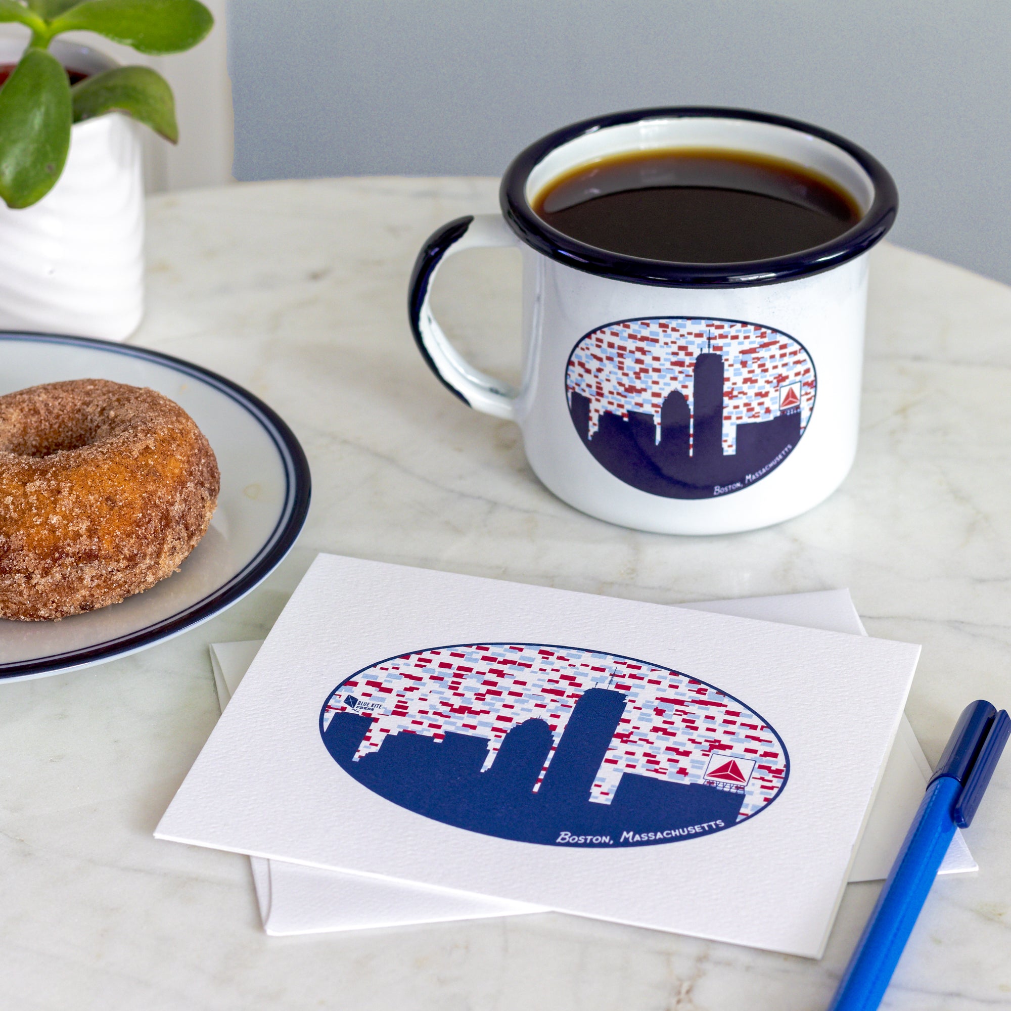 White enamel mug filled with black coffee and matching Boston skyline card on marble table with donut and pen nearby