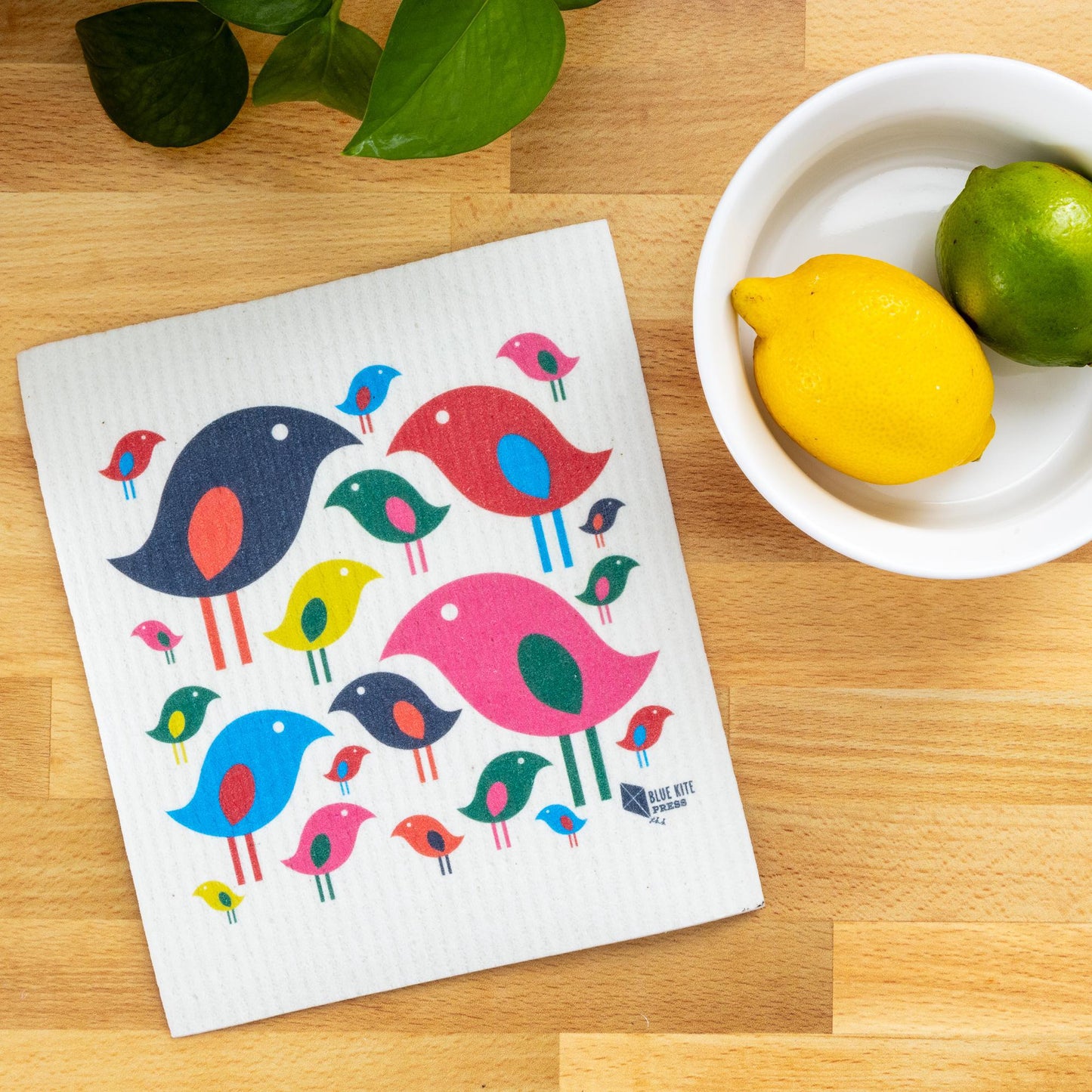 Colorful Swedish dishcloth with modern bird flock design displayed on wooden countertop next to white bowl with lemon and lime, showcasing eco-friendly kitchen cleaning alternative