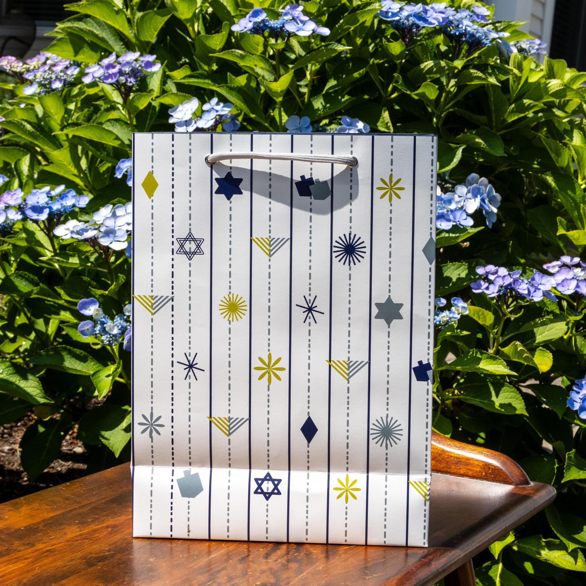 White gift bag with modern Hanukkah design featuring stylized dreidels, menorahs, Stars of David, and geometric shapes in blue, yellow, and gray, displayed on wooden surface surrounded by blue hydrangeas