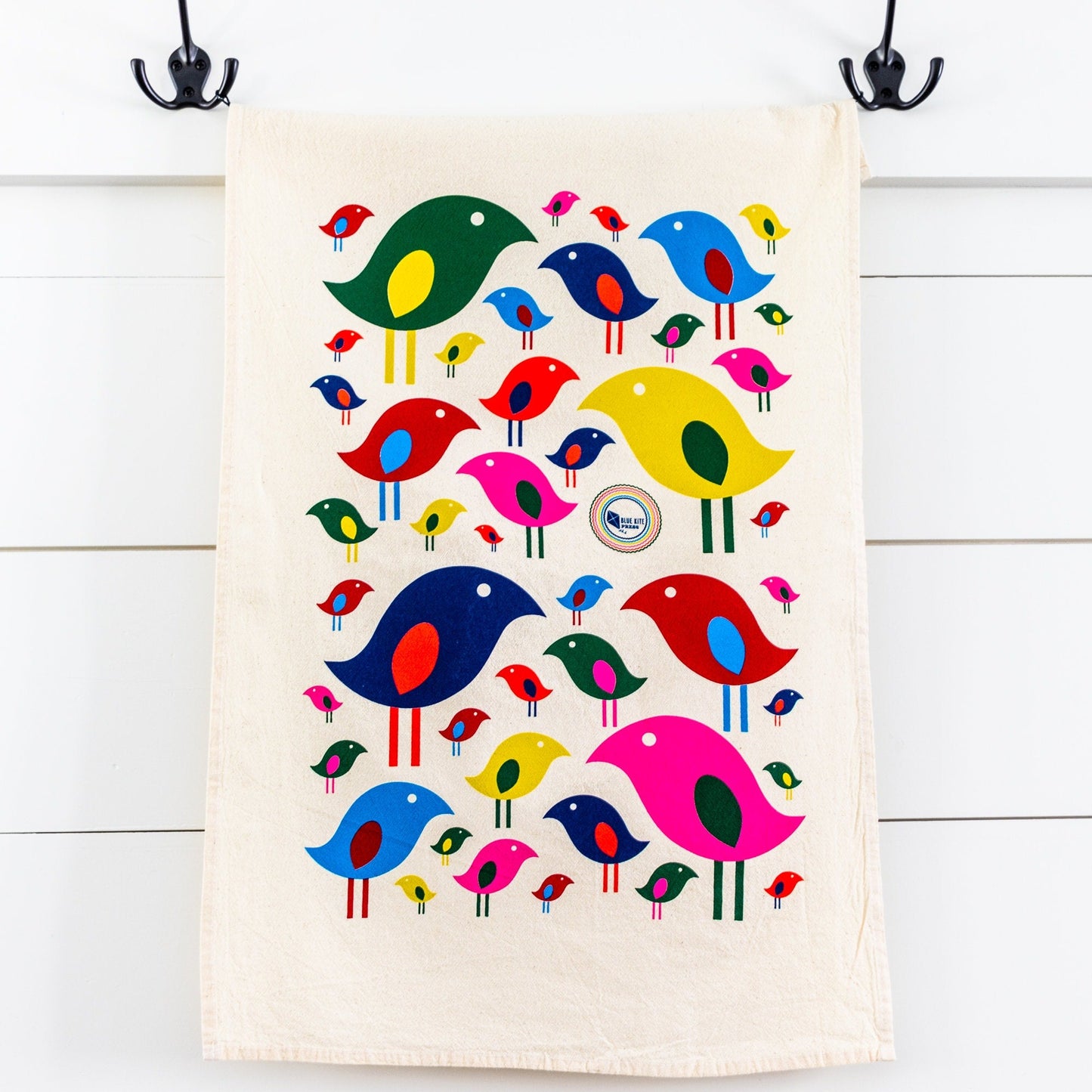 Colorful mid-century inspired bird pattern on natural cotton tea towel, featuring vibrant cartoon birds in various sizes and colors, hanging from black hooks against white wooden background