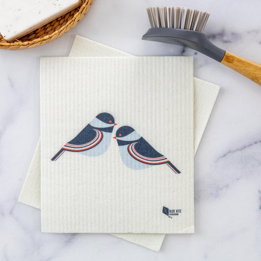 Chickadee Swedish Dishcloth featuring two stylized blue and red chickadees on a white cellulose background, shown with cleaning brush on marble surface, modern Scandinavian design for eco-friendly kitchen cleaning