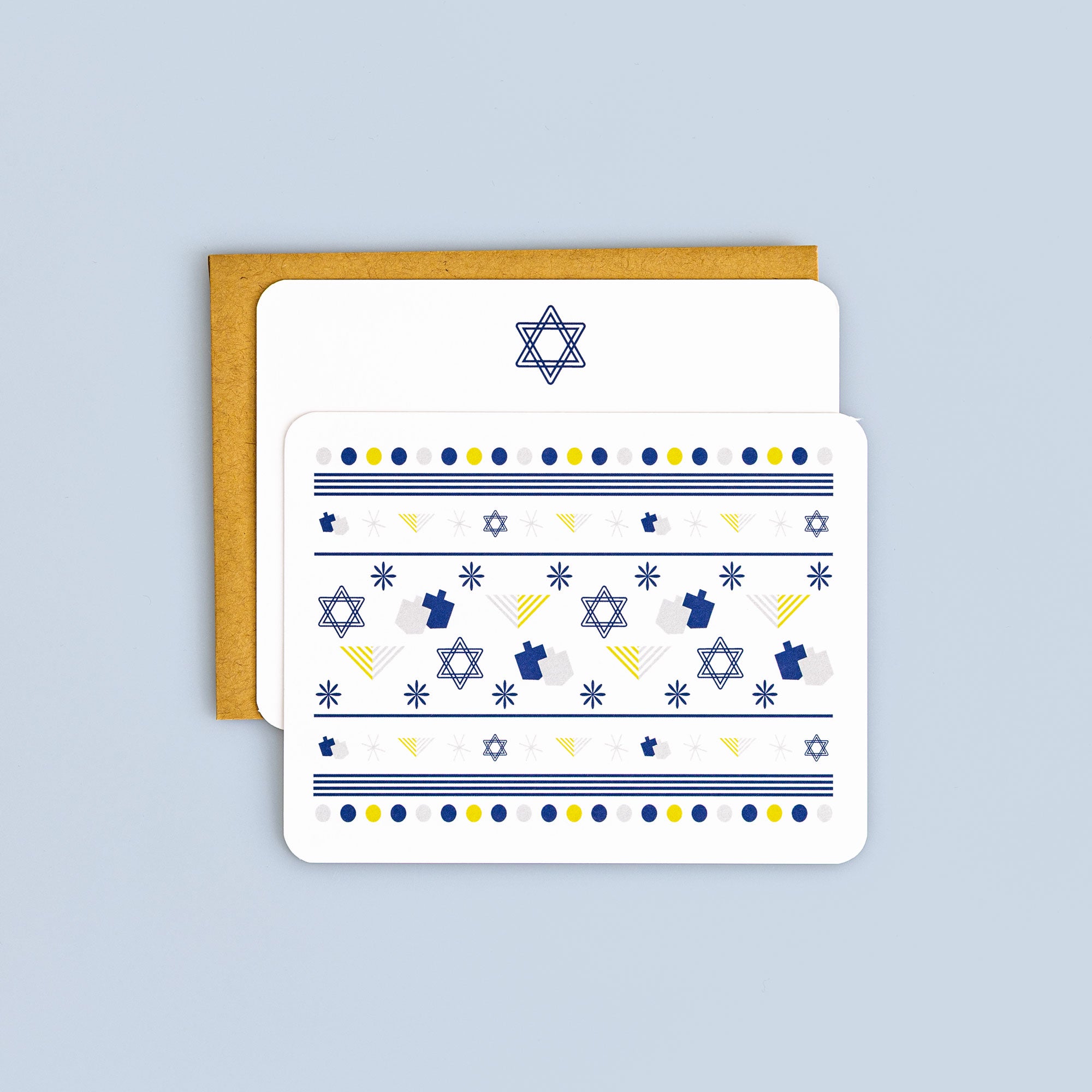 Flat Hanukkah card with blue menorahs, Stars of David, dreidels, and yellow and blue geometric patterns with kraft envelope