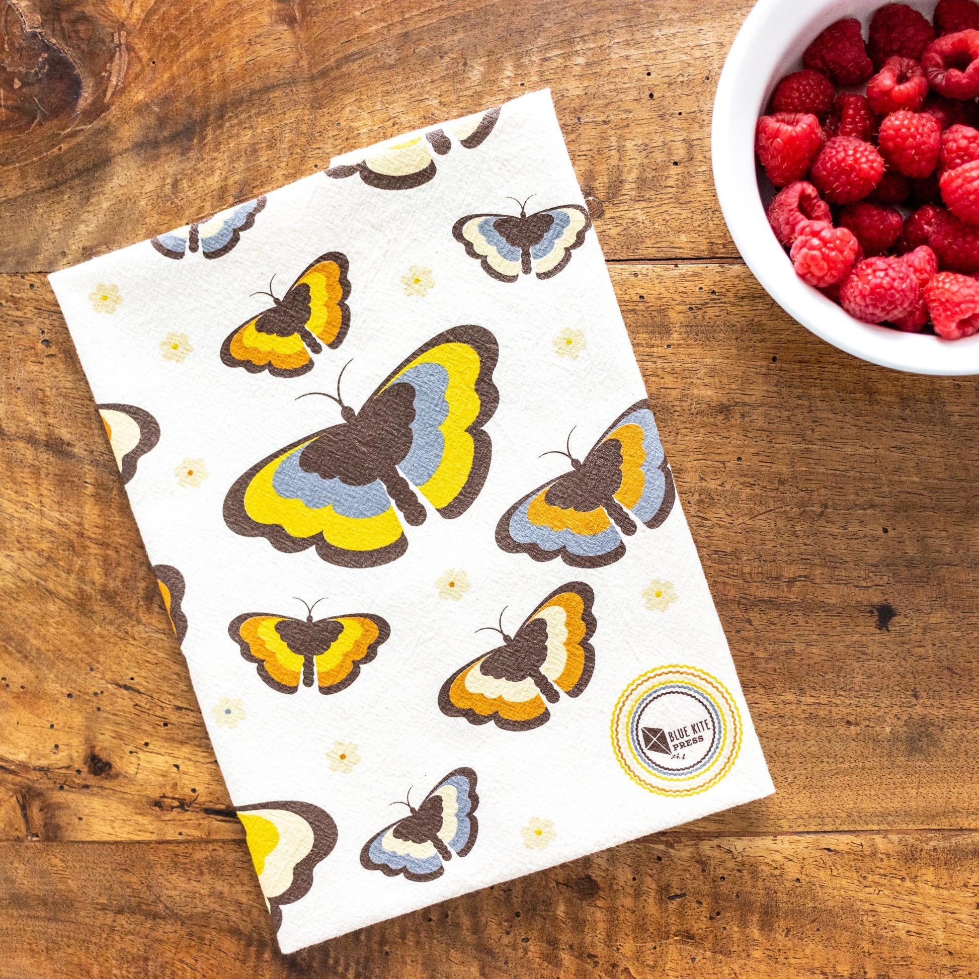 Retro butterfly patterned flour sack towel with 70s-inspired design on wooden table next to bowl of fresh raspberries, showcasing vintage kitchen aesthetic and practical use