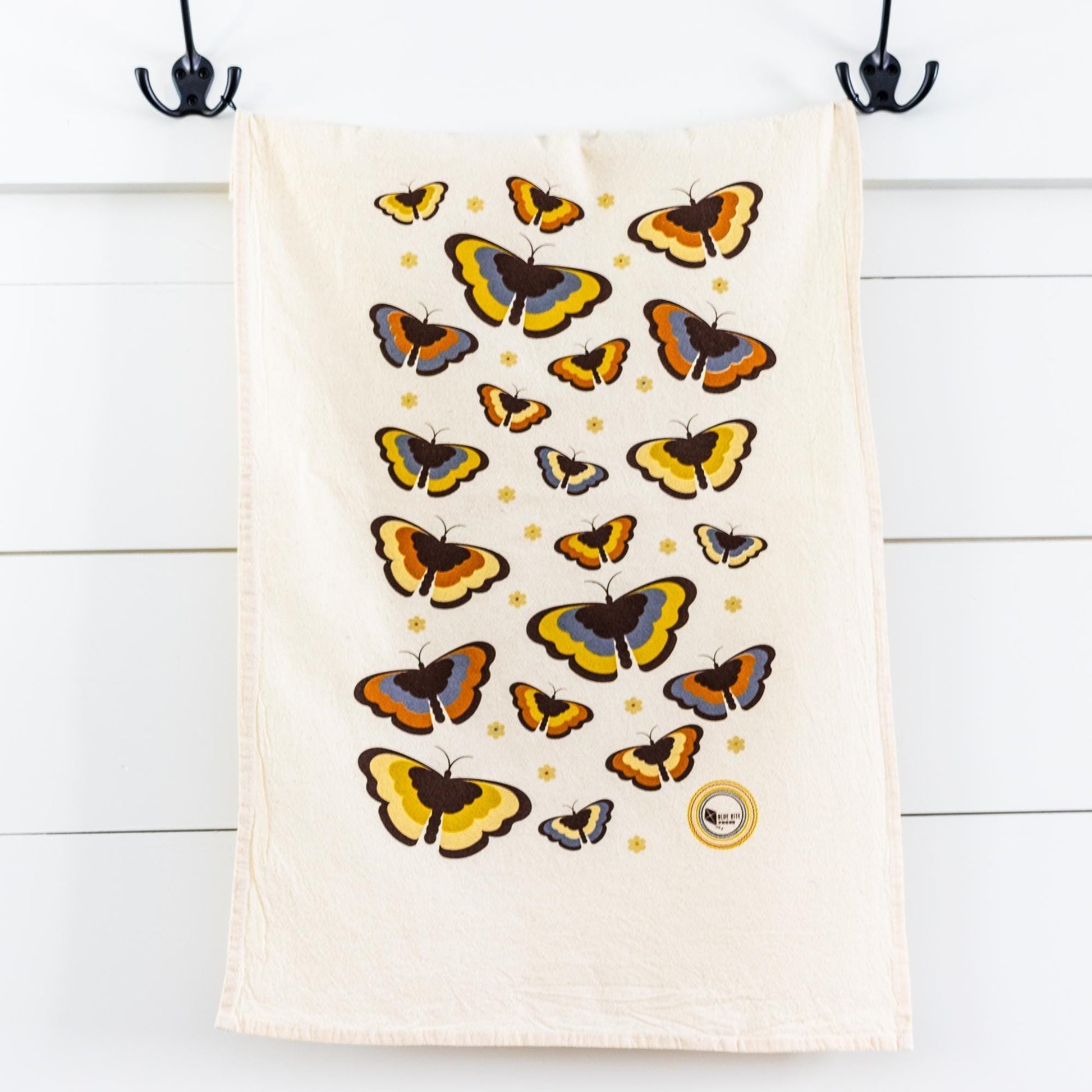 Retro-style flour sack towel with colorful butterfly pattern in yellow, orange, and blue tones, hanging on black hooks against white wooden background