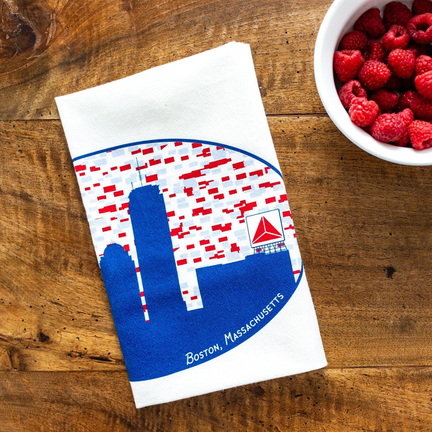 Boston skyline flour sack towel featuring blue silhouette and iconic Citgo sign, displayed on wooden surface next to bowl of fresh raspberries