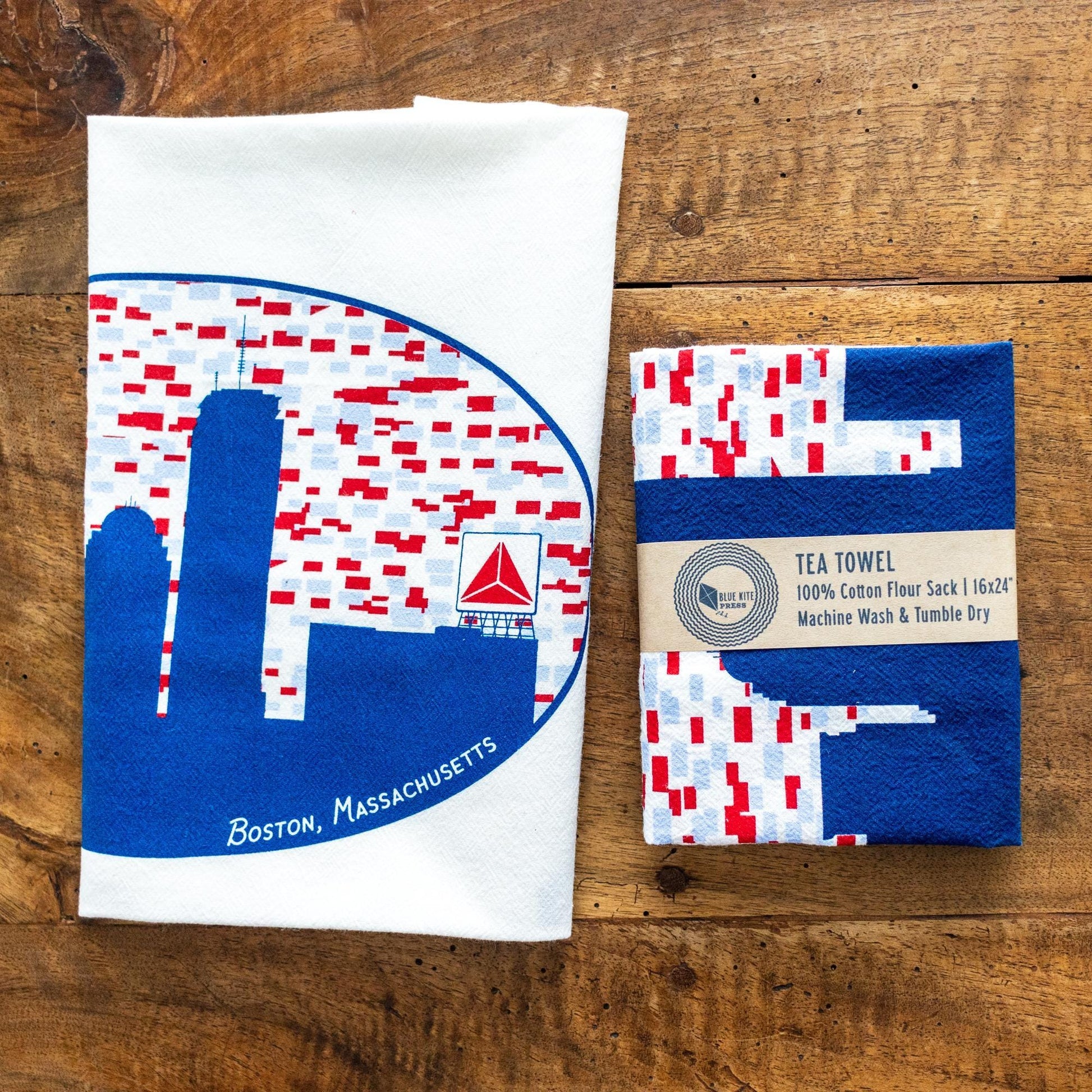 White cotton flour sack towel with blue and red Boston skyline design featuring iconic Citgo sign, displayed next to matching packaging on wooden surface