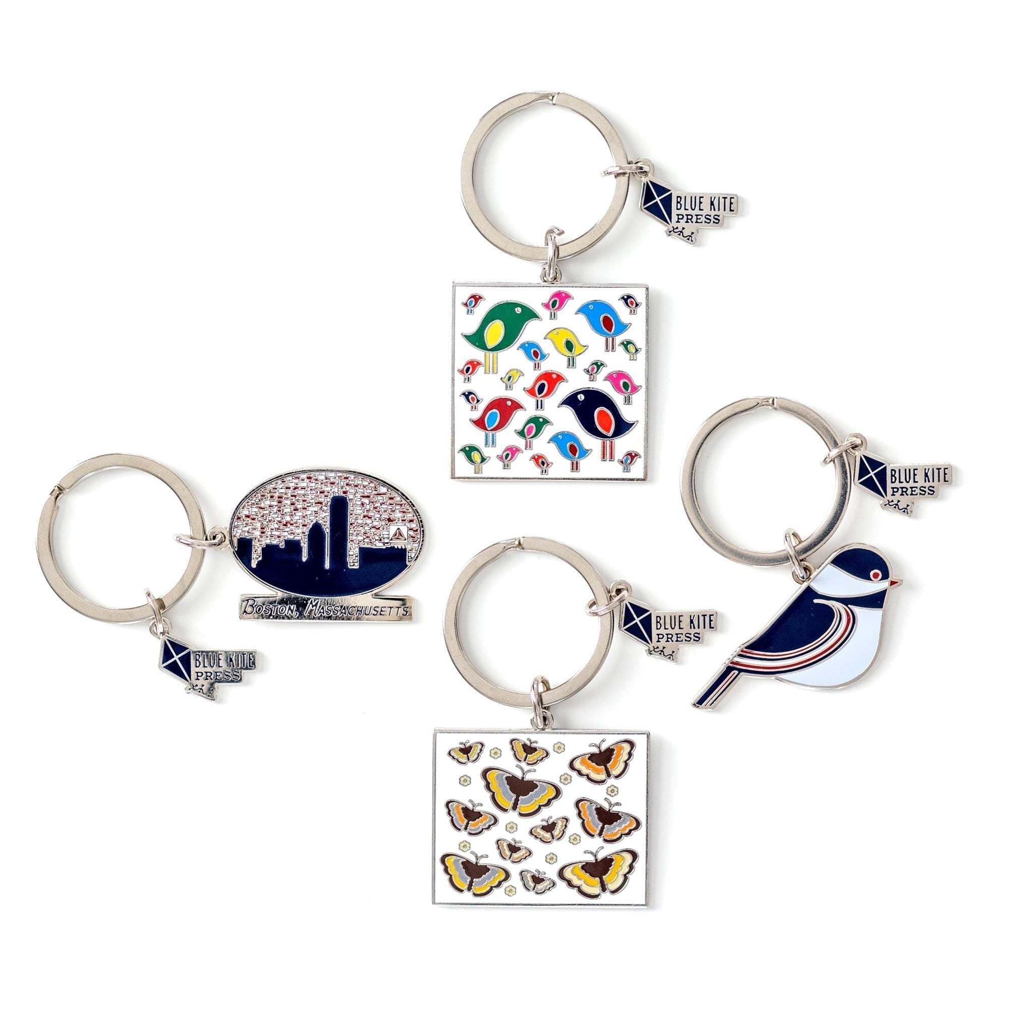 Colorful enamel keychains featuring retro designs including butterflies, birds, and cityscape. Silver rings with Blue Kite brand tags attached. Square pendants with vibrant patterns in 70s-inspired color palette.