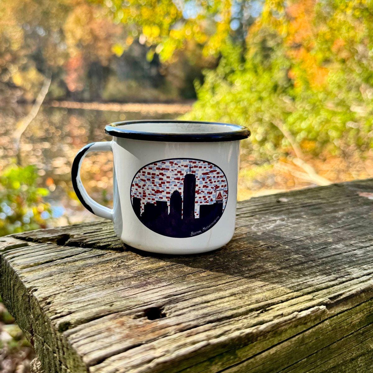 White enamel mug with Boston skyline design featuring Citgo sign on wooden surface outdoors, surrounded by colorful autumn foliage in background