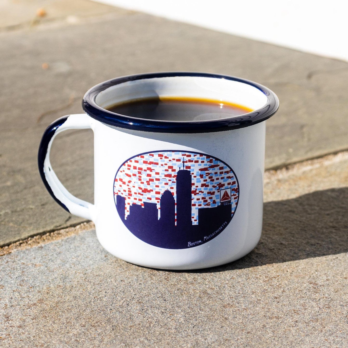 White enamel mug with Boston skyline design featuring Citgo sign, filled with coffee, on concrete surface. Navy blue rim and handle, stylized city silhouette with red accents representing lights or windows.