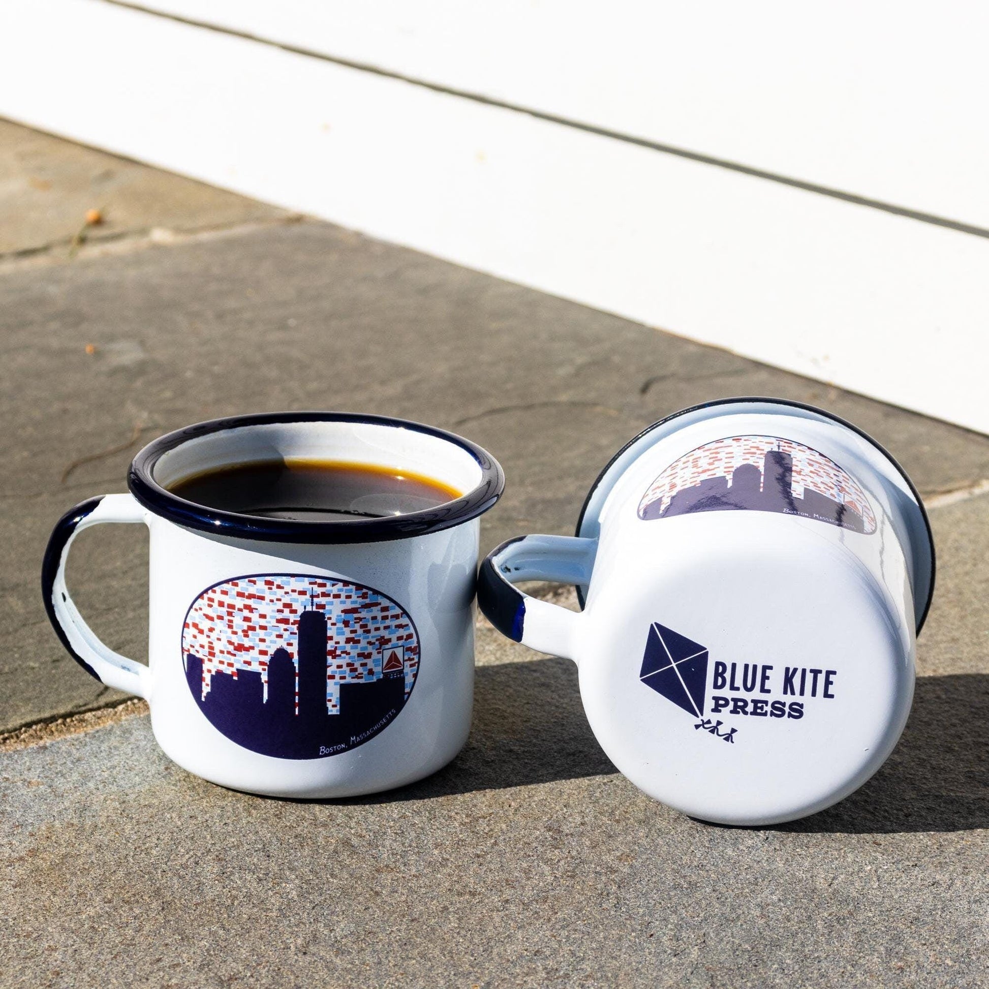 White enamel mugs with Boston skyline design featuring Citgo sign, one filled with coffee, on stone surface. Blue Kite Press logo visible on mug bottom.