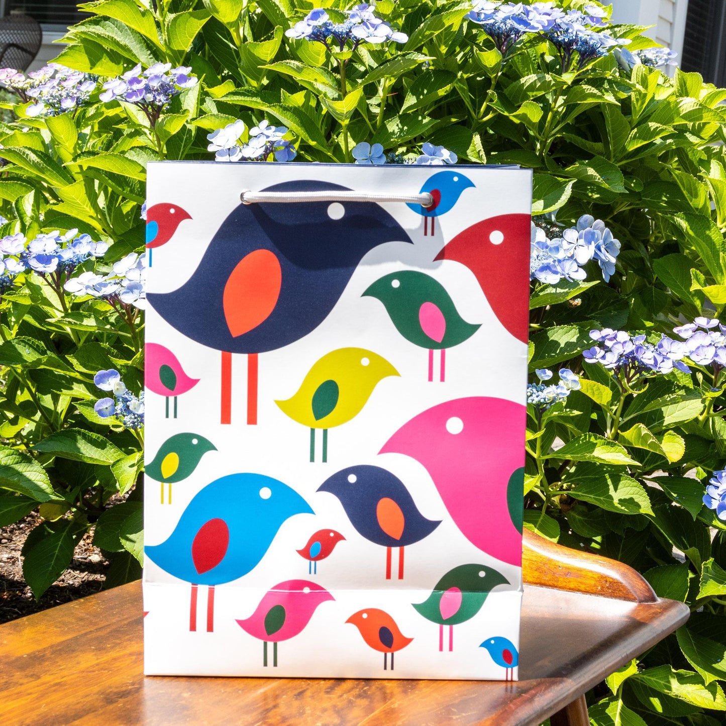 White gift bag with colorful modern bird flock design featuring stylized birds in various sizes and bright colors, displayed outdoors against green foliage with blue flowers