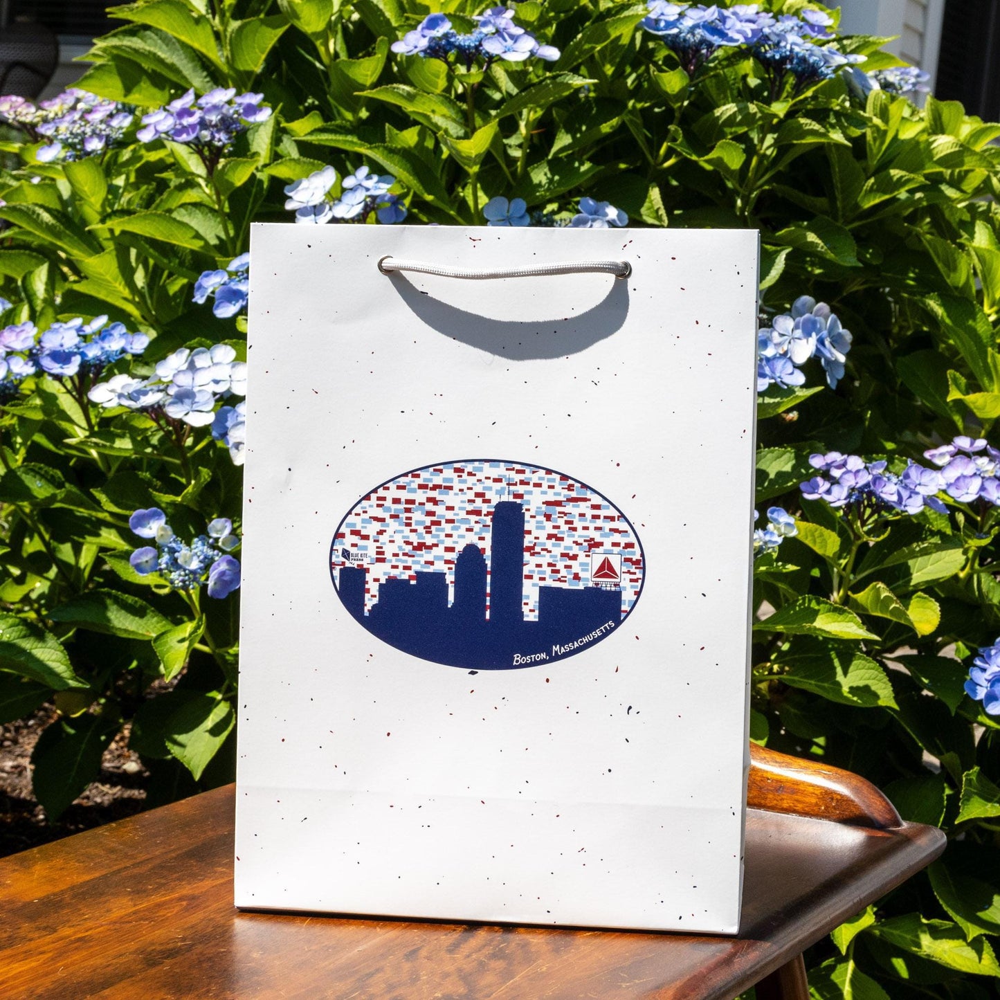 White gift bag with Boston skyline silhouette featuring Citgo sign, surrounded by blue hydrangeas on wooden surface, perfect for presenting Boston-themed gifts