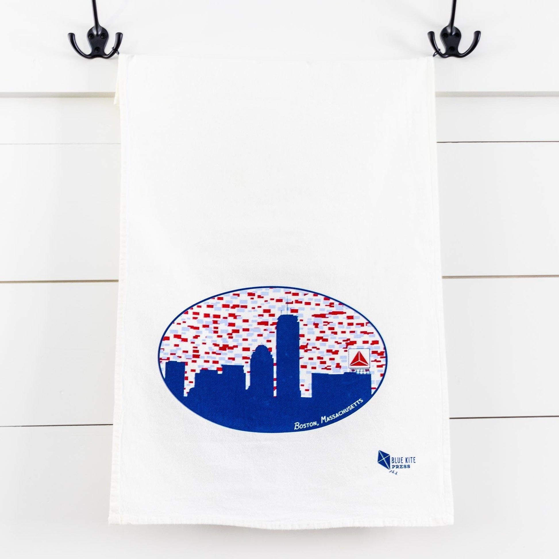 White cotton flour sack towel hanging from hooks, featuring blue Boston skyline silhouette with iconic Citgo sign in oval shape, dotted with red accents, perfect for kitchen decor or drying dishes