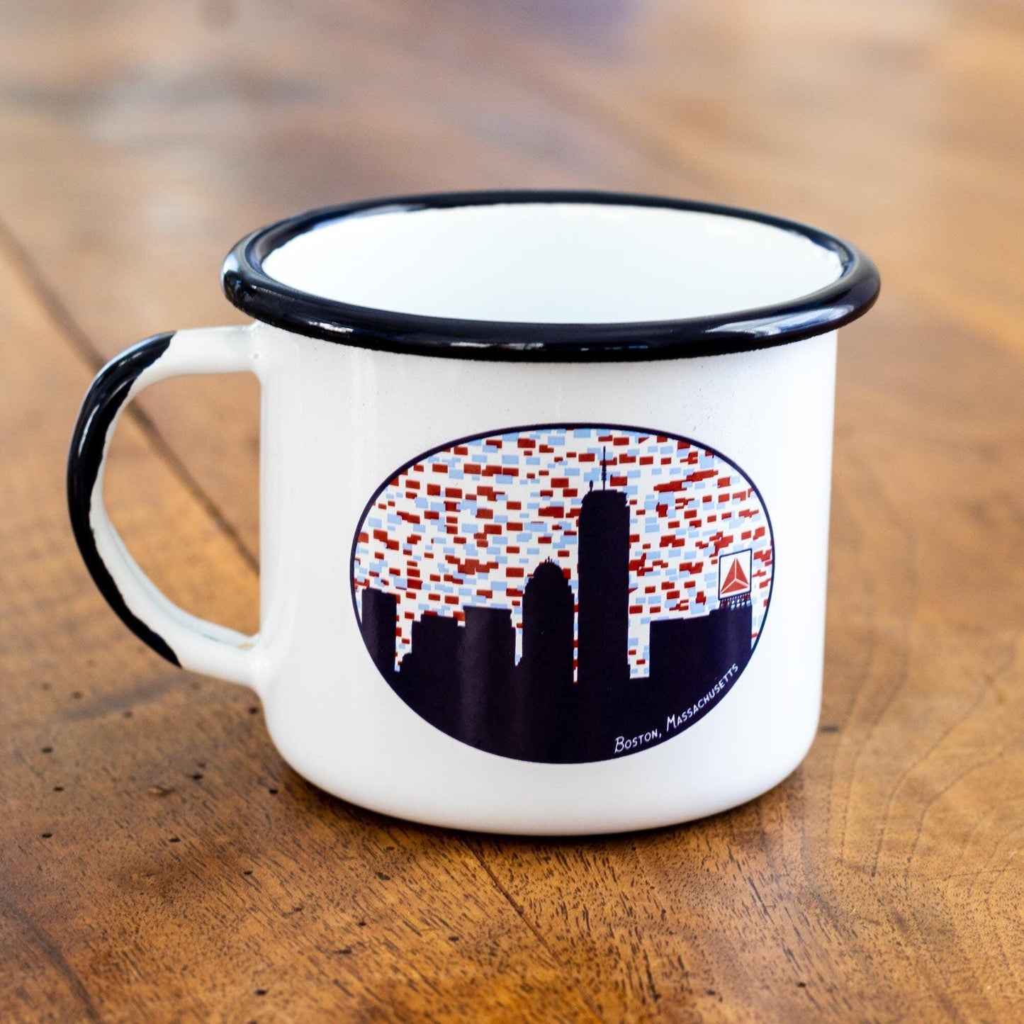 White enamel mug with black rim featuring Boston skyline silhouette and iconic Citgo sign design, set on wooden surface. Perfect for coffee or tea lovers and Boston enthusiasts.