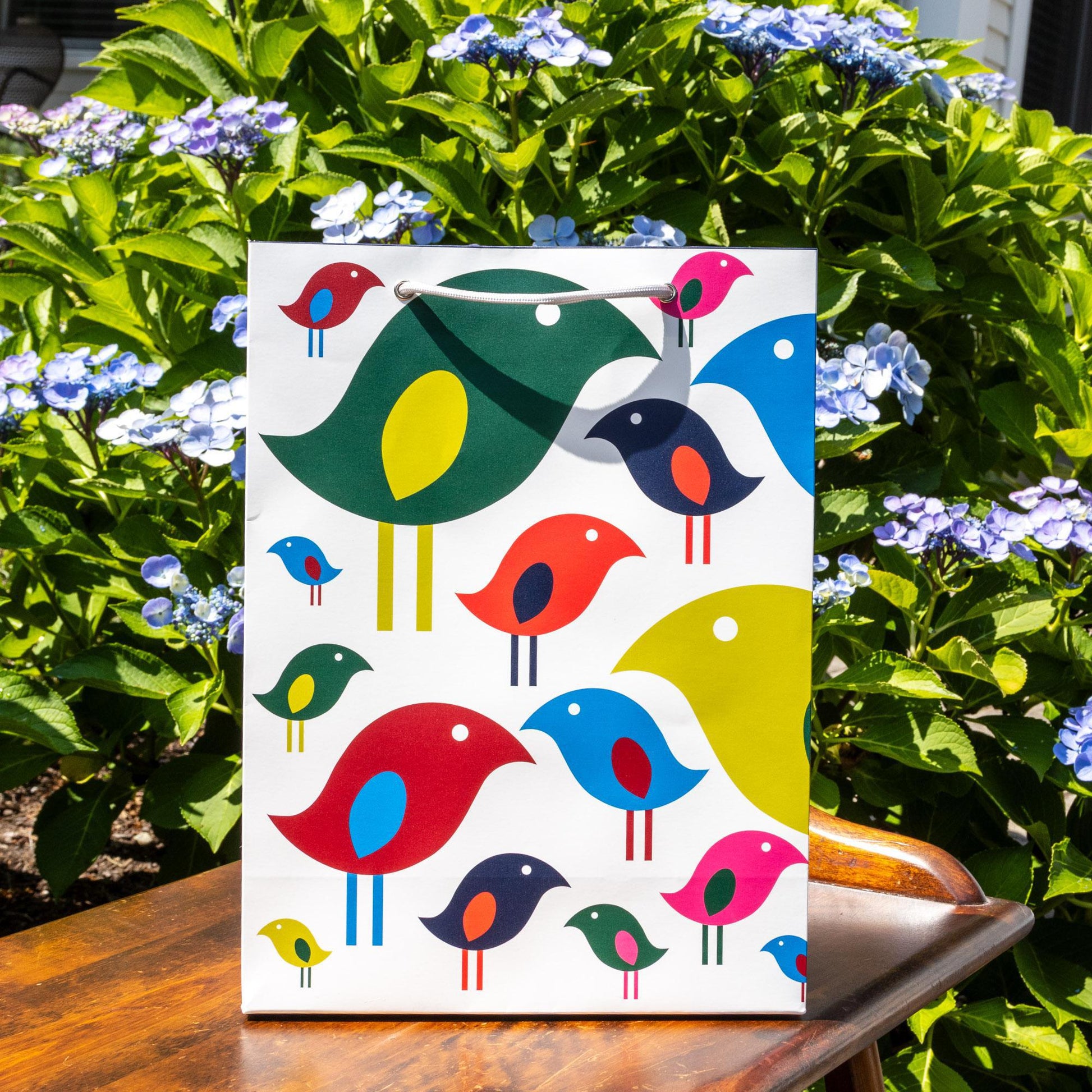 Colorful gift bag featuring modern, stylized bird design with various bright hues against white background, displayed outdoors amid green foliage and blue flowers