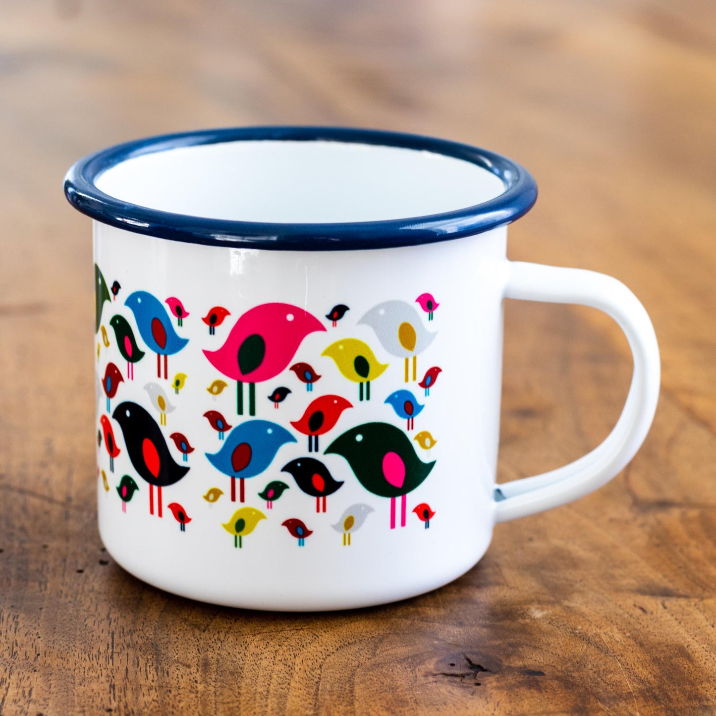 Bright Bird Enamel Mug featuring a playful pattern of colorful cartoon birds in red, blue, black, and yellow on white enamel, with navy blue rim. Durable 12 oz camping-style mug displayed on wooden surface.