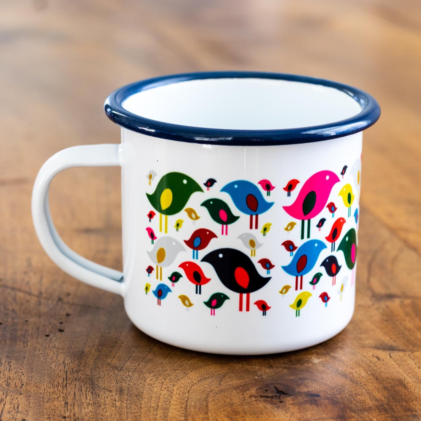 Bright Bird Enamel Mug featuring colorful cartoon birds in green, blue, pink, red, and black on white enamel, with navy blue rim. 12-ounce camping-style mug photographed on wooden surface.