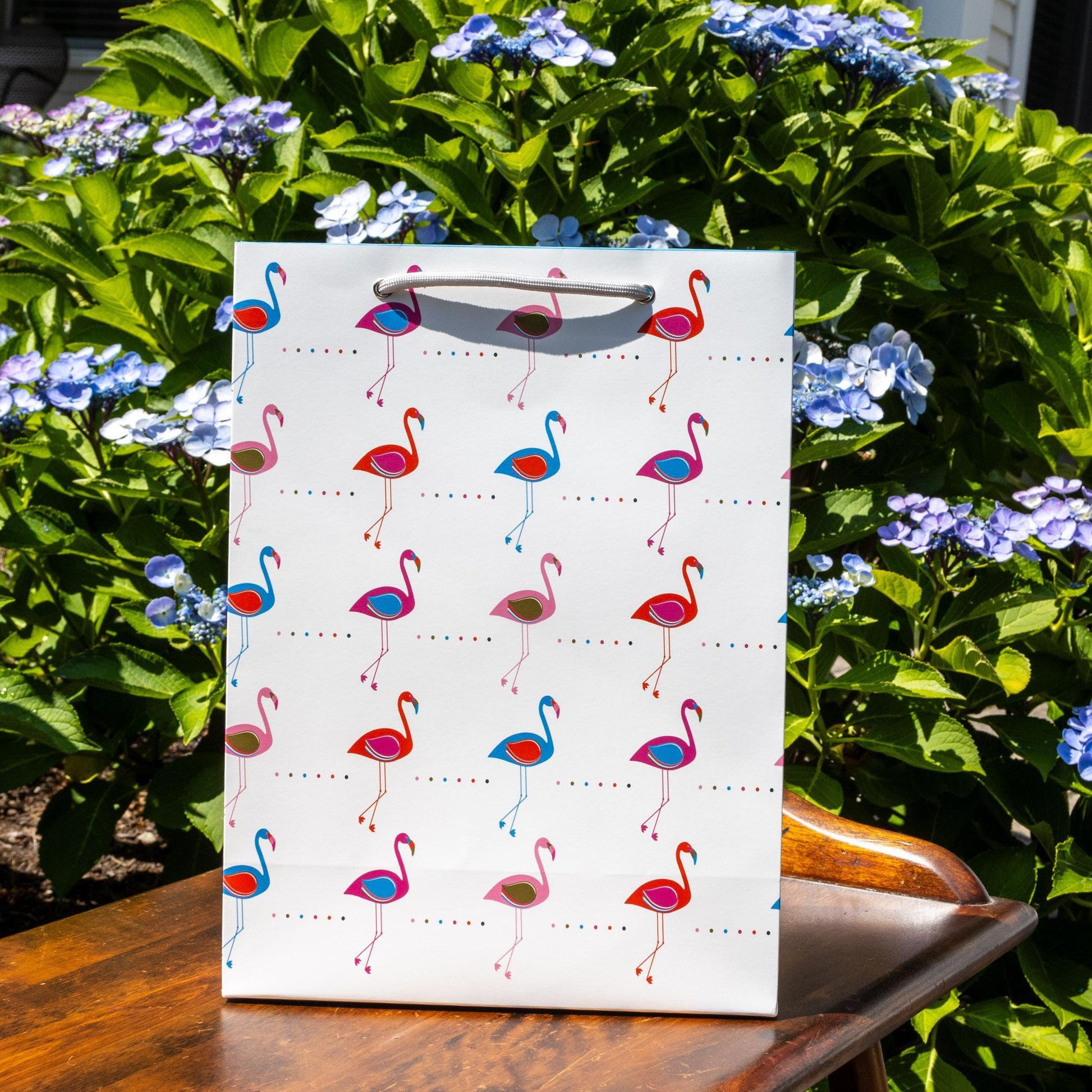 Modern gift bag featuring colorful flamingo pattern on white background, displayed outdoors surrounded by blue hydrangea flowers and green foliage