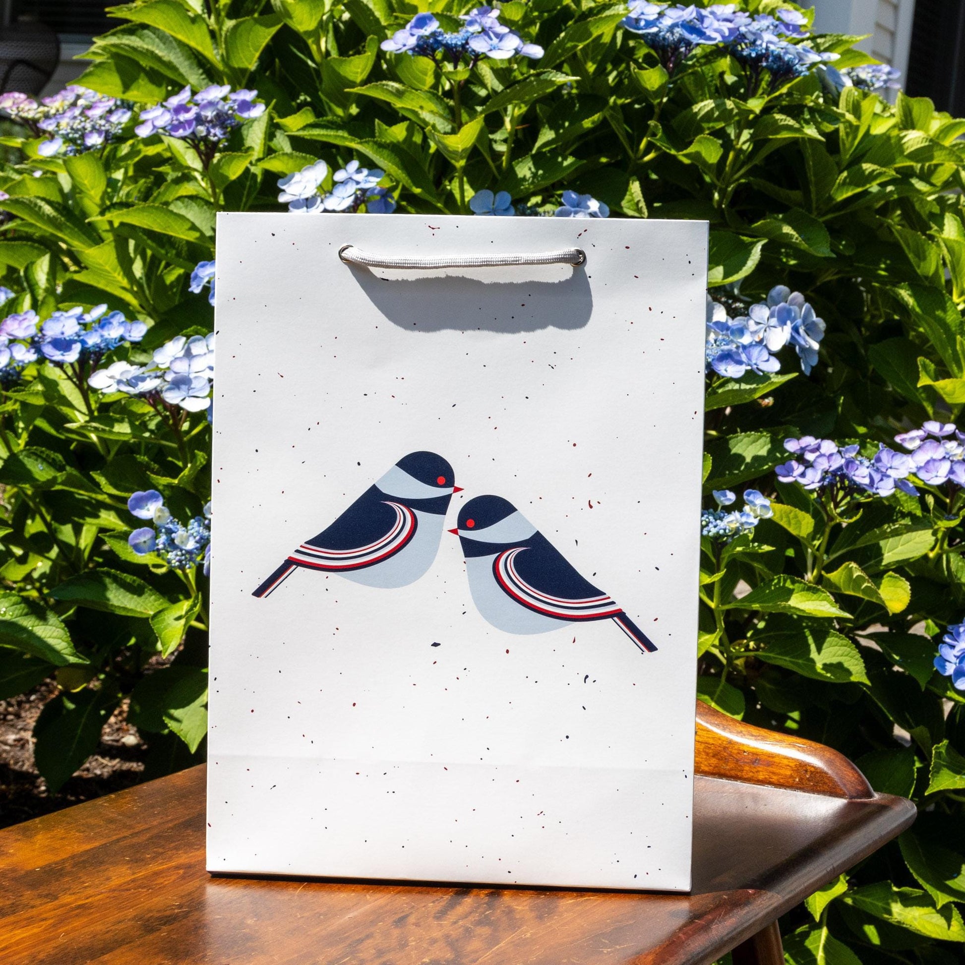 White matte paper gift bag with modern chickadee design, featuring two stylized birds in navy and red, displayed outdoors against vibrant blue hydrangea flowers and green foliage