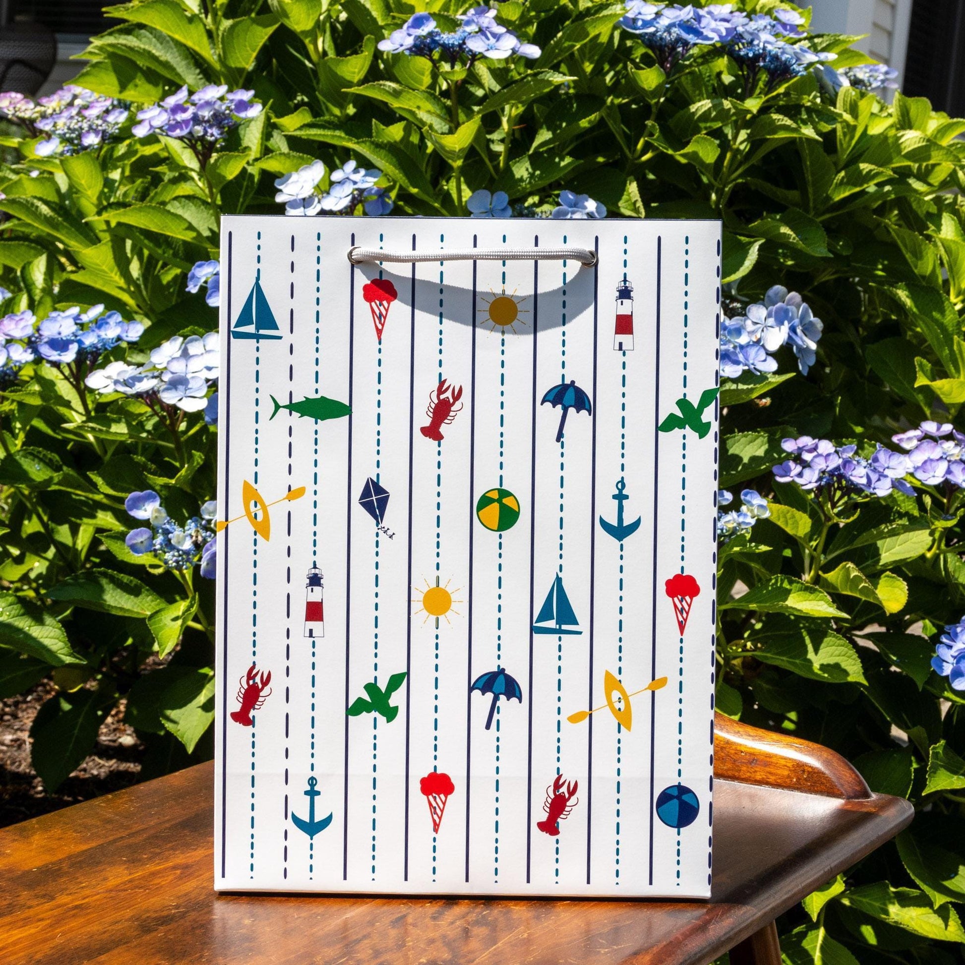 White gift bag with colorful New England coastal motifs including sailboats, lighthouses, fish, anchors, and buoys, surrounded by vibrant blue hydrangea flowers and lush green leaves