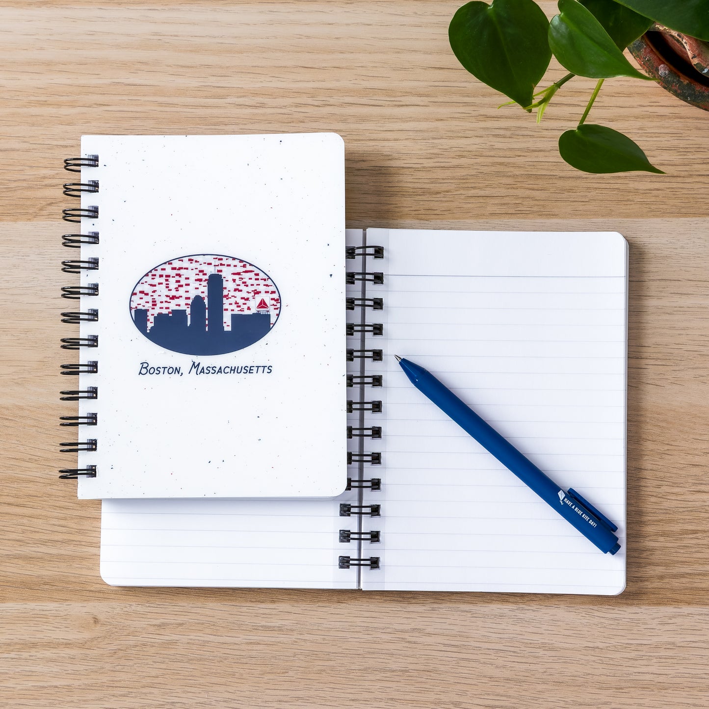 Spiral-bound notebook featuring Boston skyline with Citgo sign on cover, open to blank page with blue pen, on wooden surface with green plant leaves