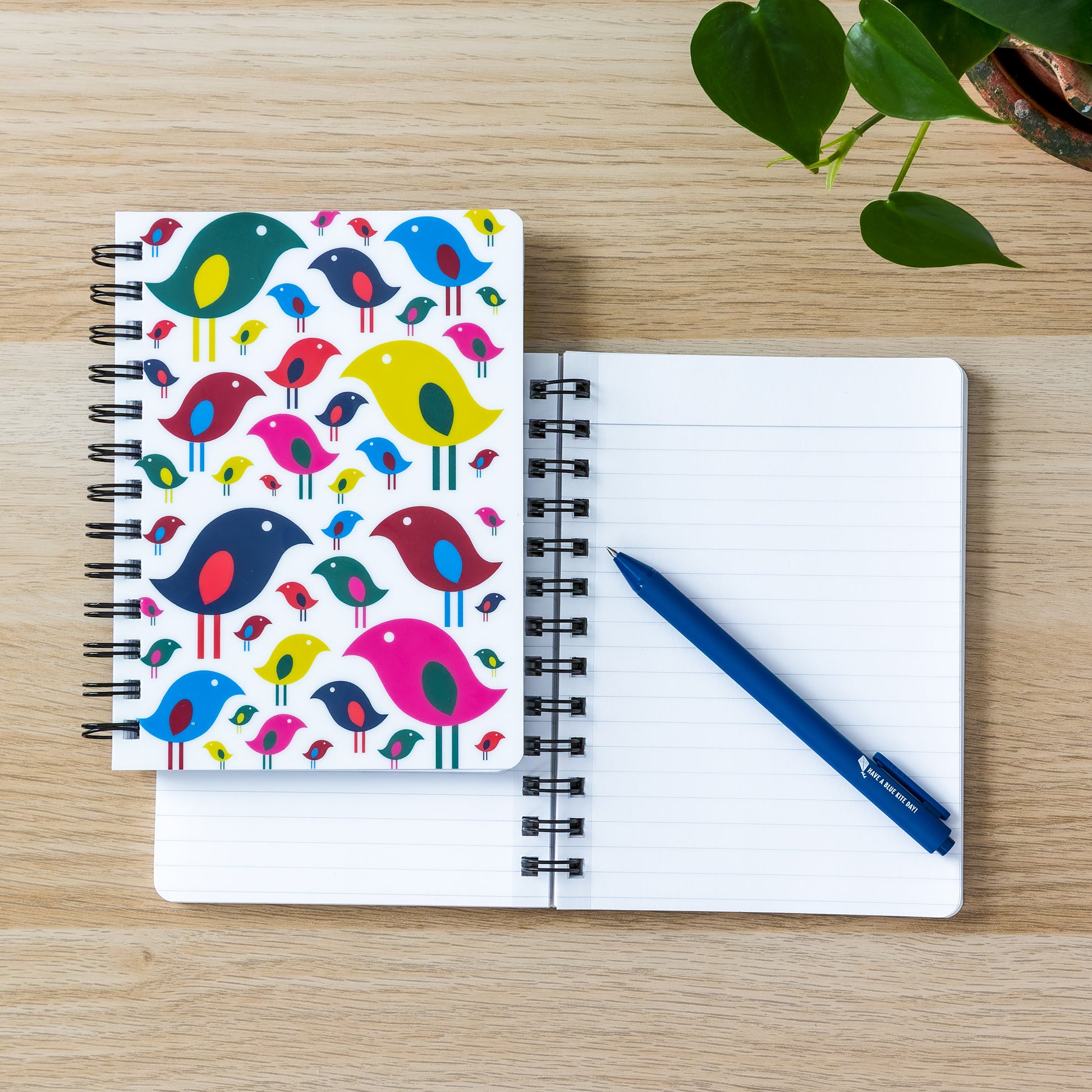 Colorful bird flock design spiral notebook with blank pages and blue pen on wooden surface, next to green plant leaves