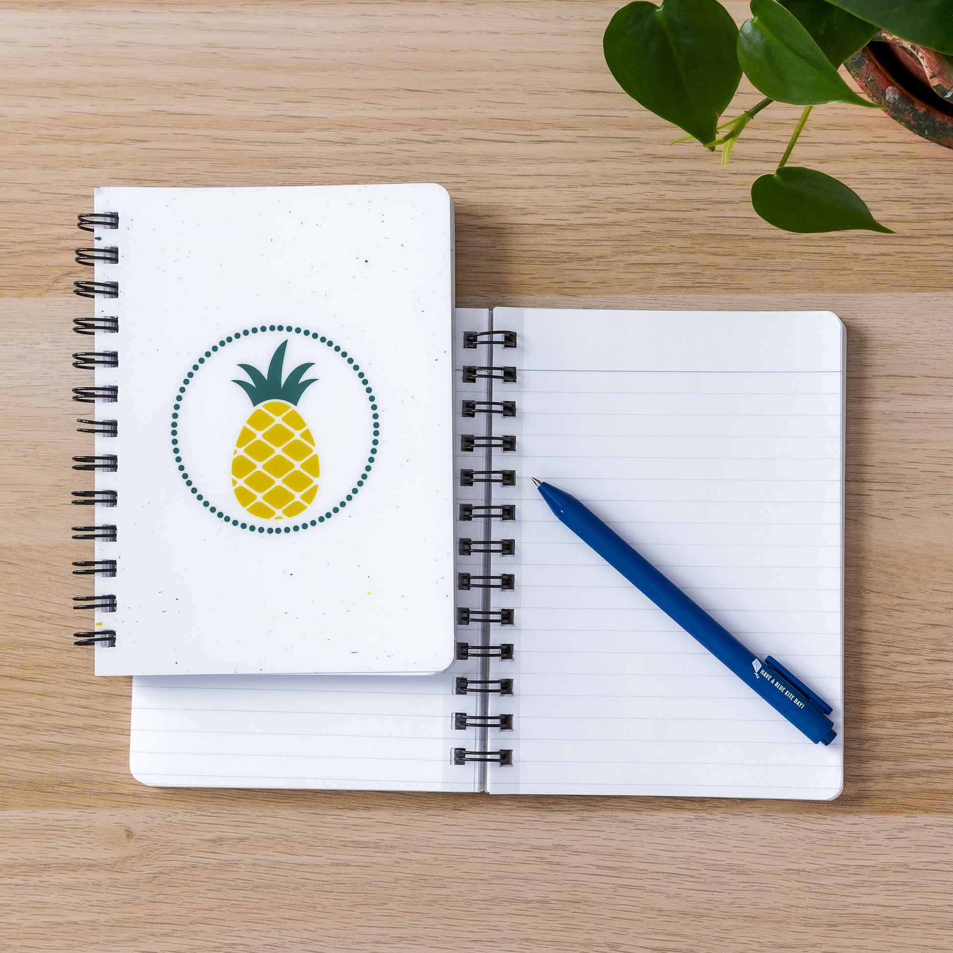 Spiral-bound notebooks with pineapple design on wooden surface, blue pen, and green leaves. One notebook open to blank page, the other closed showing yellow pineapple illustration on white cover.