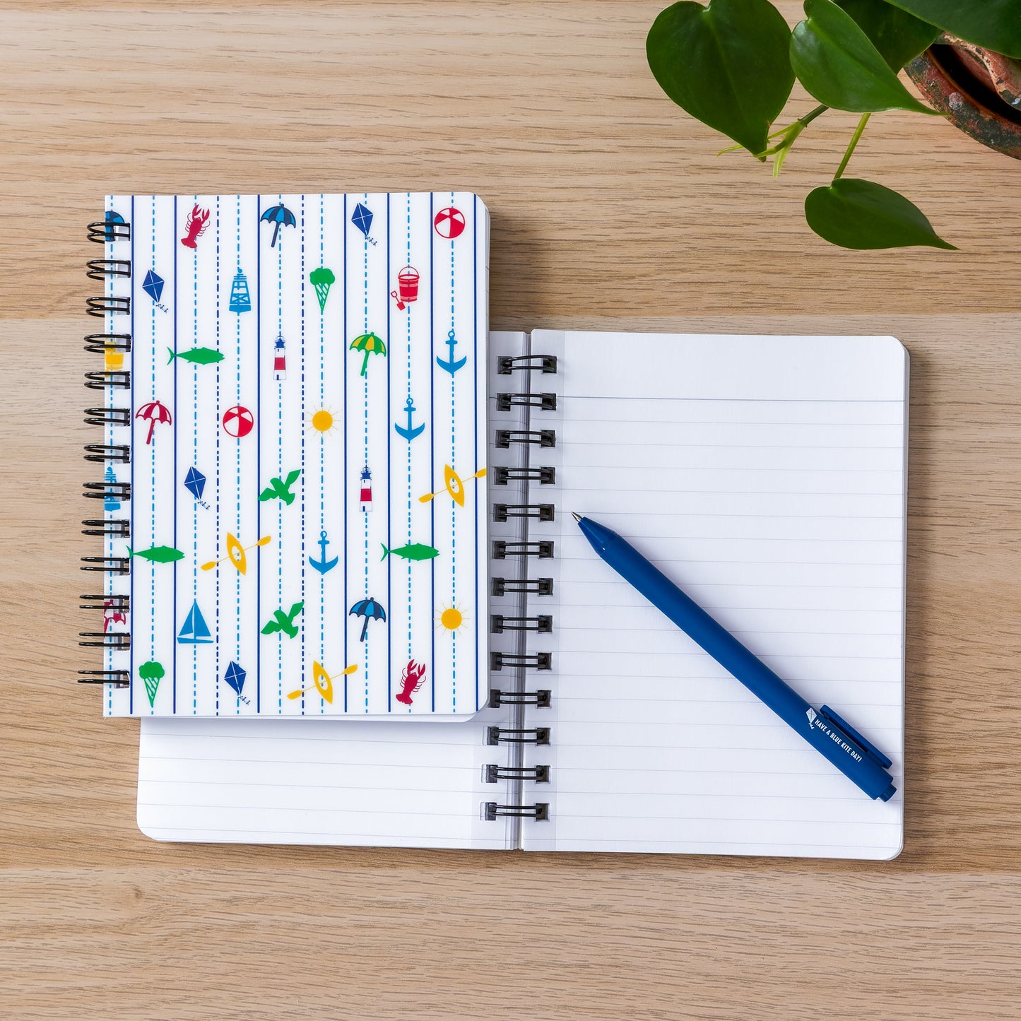 Spiral-bound notebook with colorful New England coastal icons on cover, open blank notebook, and blue pen on wooden surface with green plant leaves