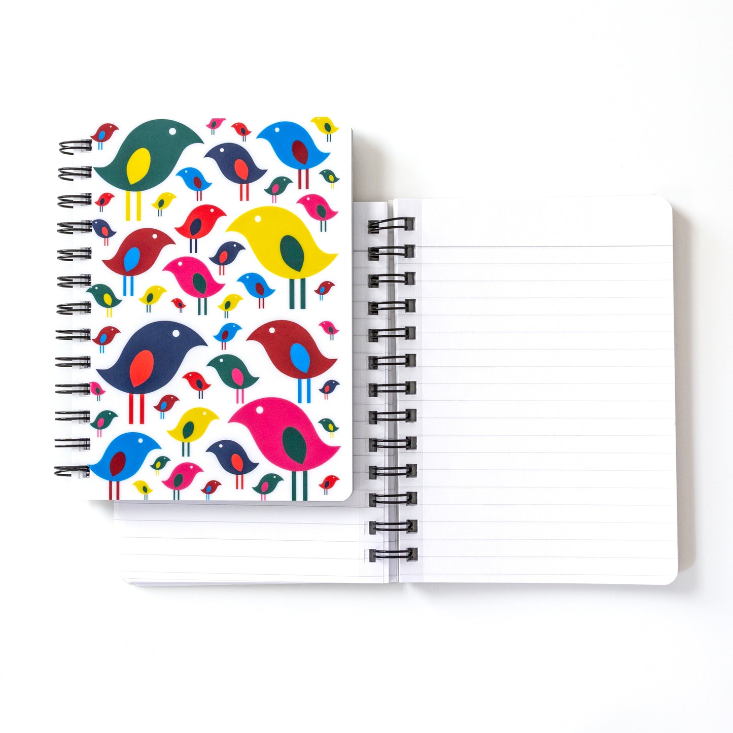 Colorful spiral-bound notebook featuring modern, stylized bird flock design on white cover. Various sizes and shapes of birds in bright colors like blue, red, yellow, and pink. Open notebook with blank pages beside it.