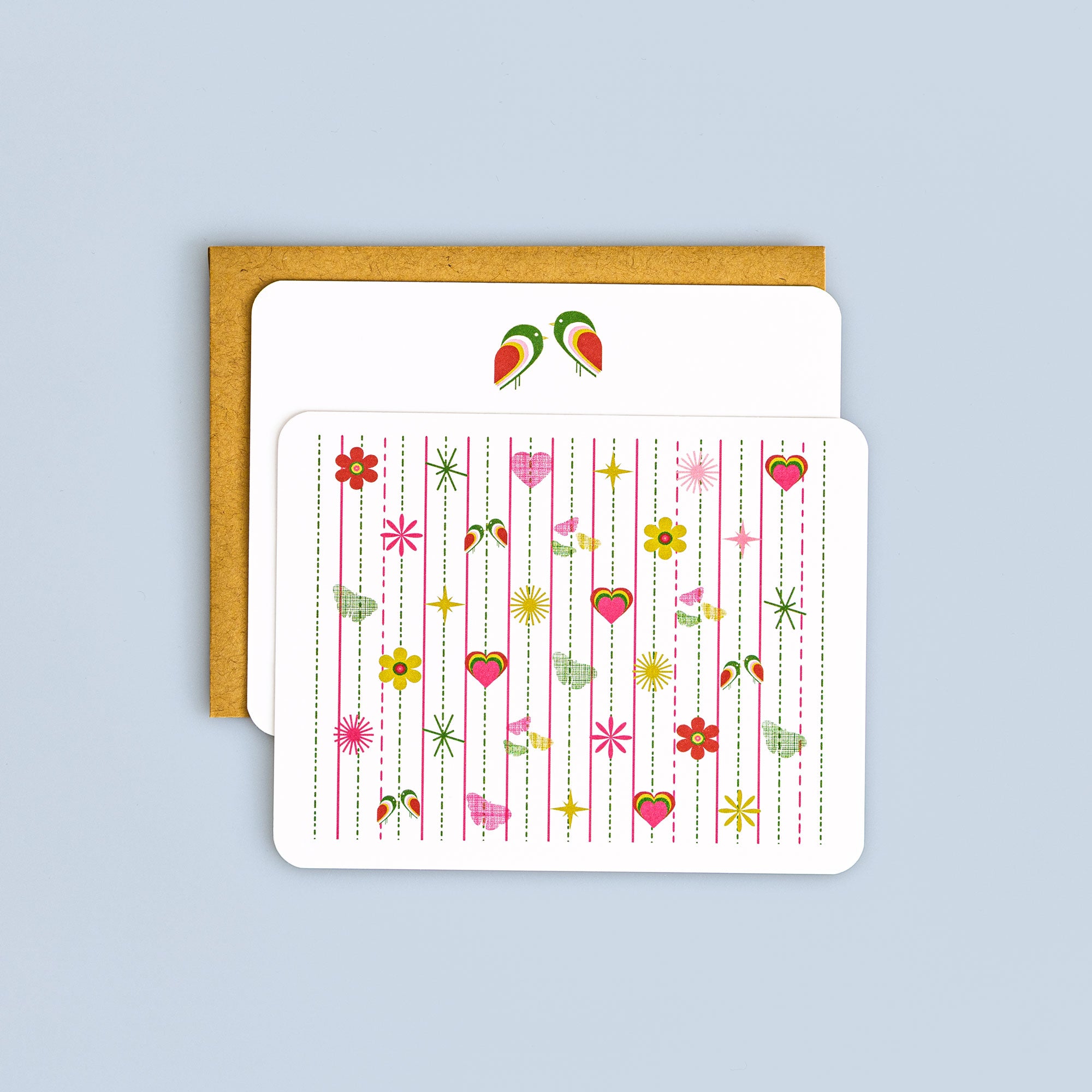 Flat pink tiki greeting card with colorful birds, hearts, and floral patterns paired with a kraft envelope on light blue background