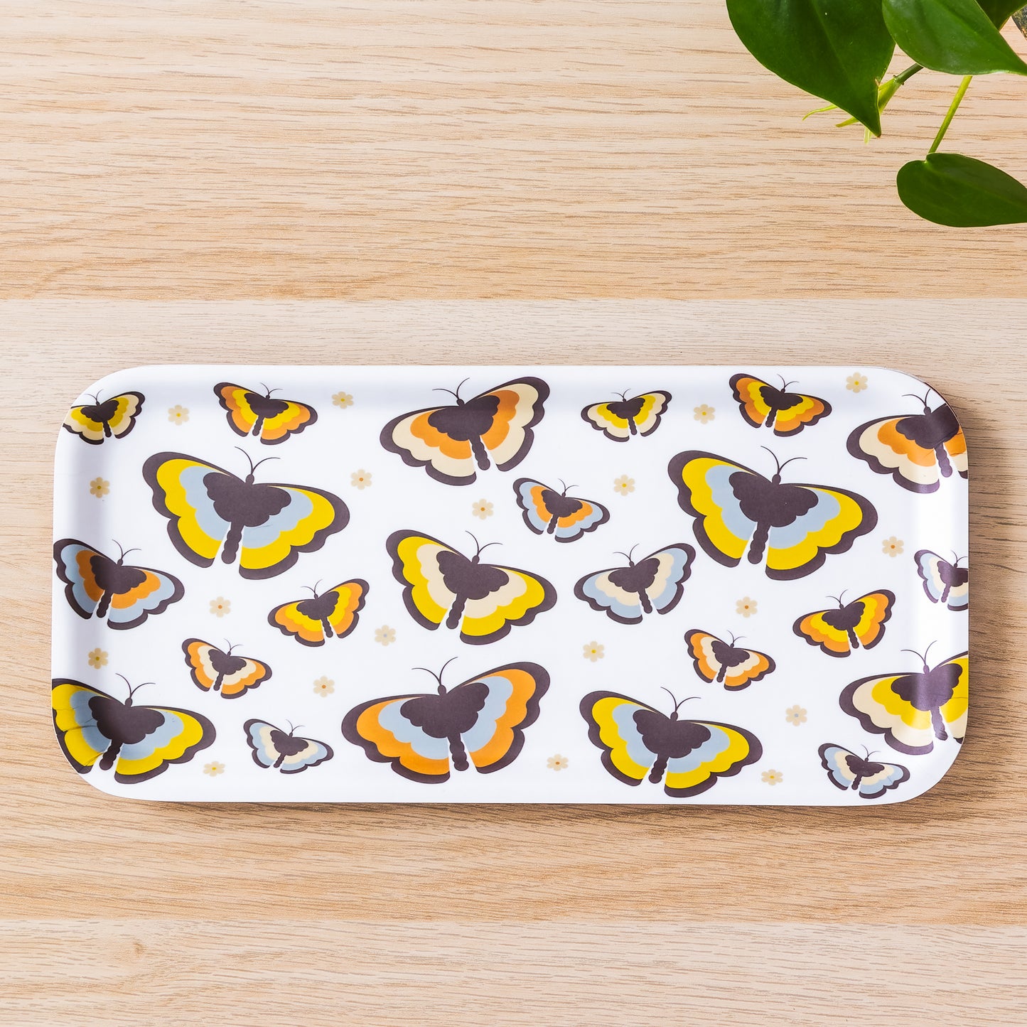 Retro 70s-style butterfly birch tray with colorful yellow and orange butterfly pattern on white background, displayed on wooden surface with green plant leaves visible