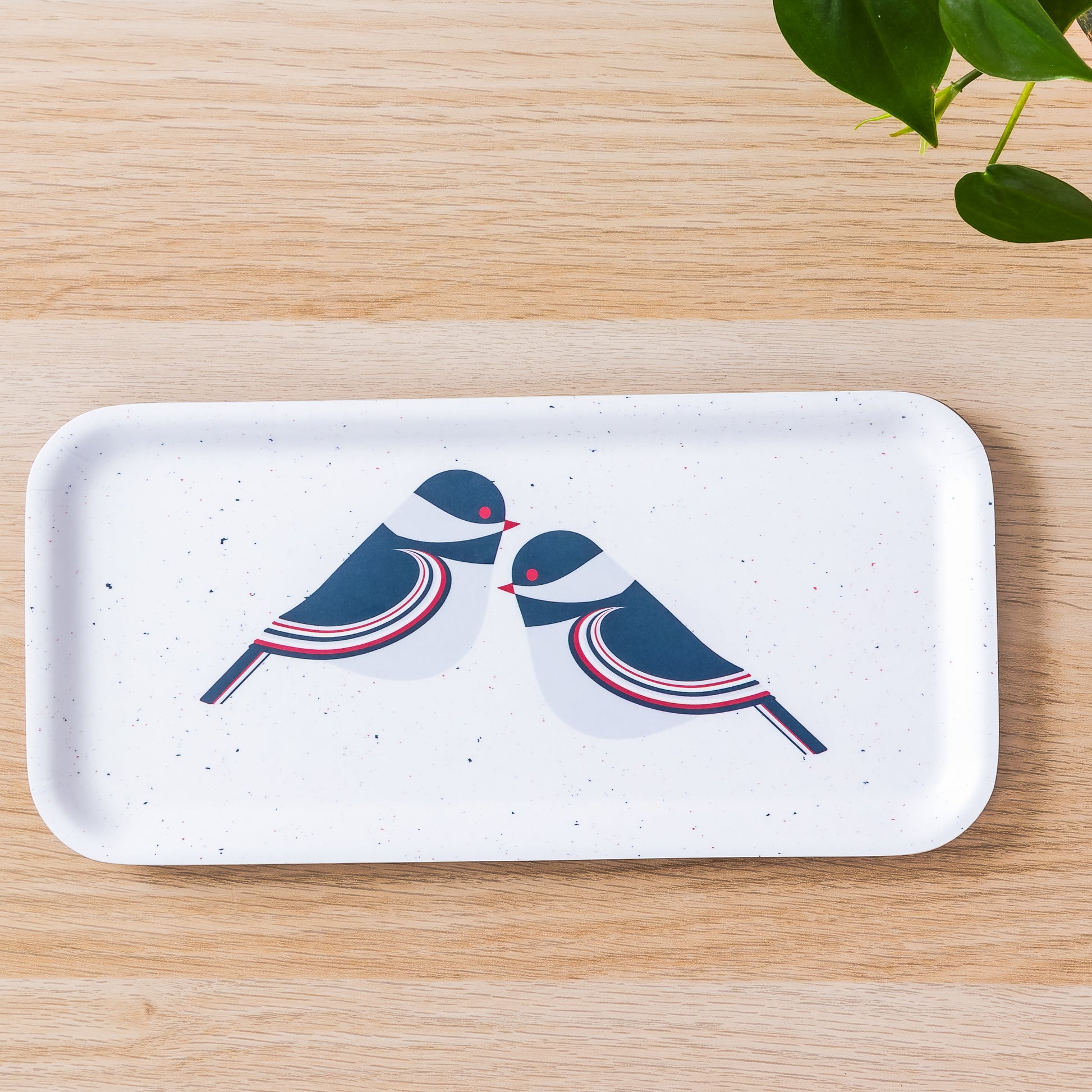 Modern white birch tray with stylized blue and red chickadee design, rectangular shape on wooden surface with green plant leaves visible